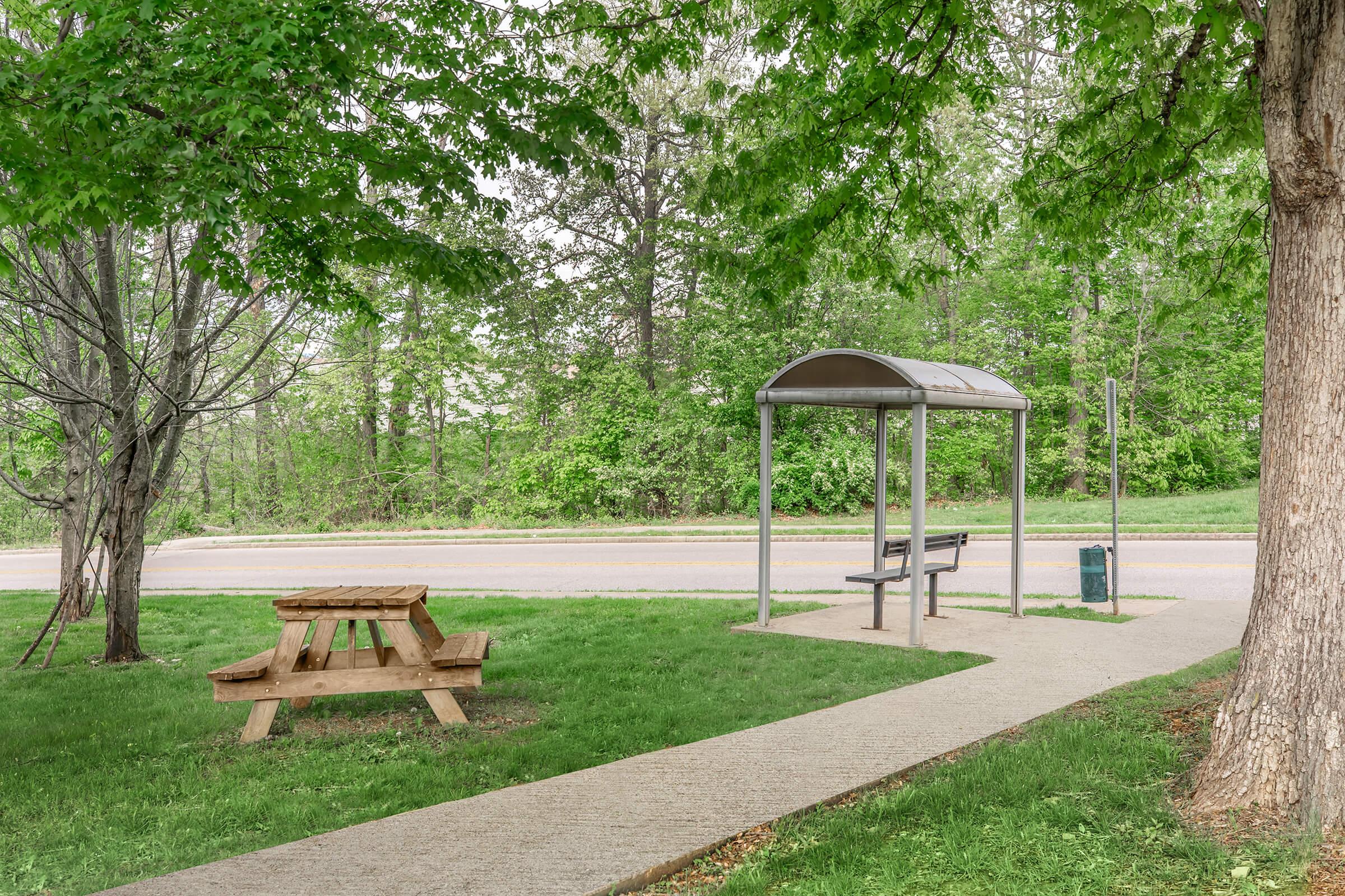 A bus stop shelter with a bench is situated along a paved path next to a road. Nearby, there is a wooden picnic table on a grassy area under trees, providing a peaceful setting. Lush greenery surrounds the scene, creating a calm and inviting atmosphere.