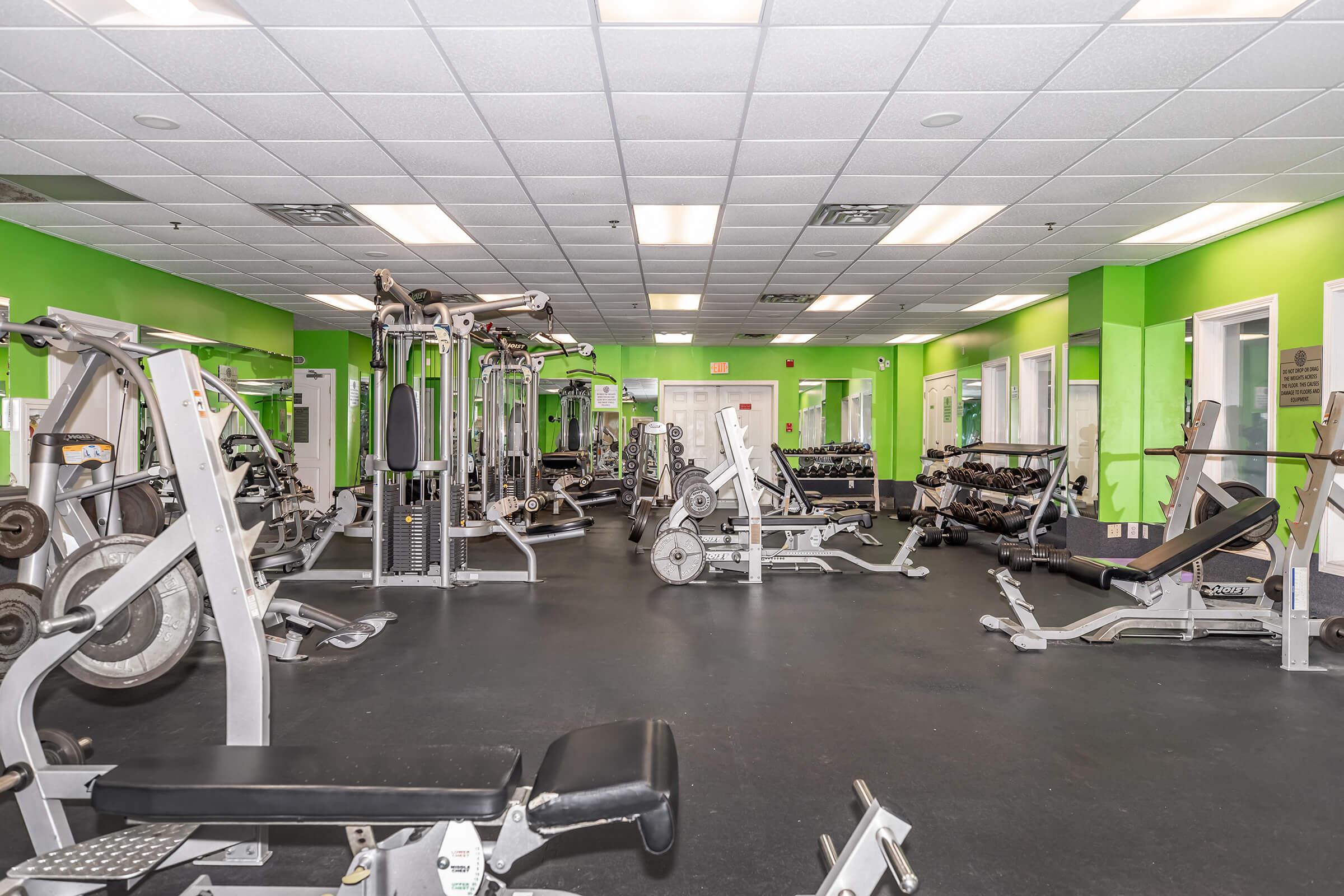 A spacious gym interior featuring various exercise machines and weightlifting equipment. The walls are painted bright green, and large mirrors are positioned throughout the room. The flooring is dark, enhancing the modern look of the fitness area, which is well-lit with overhead lighting.