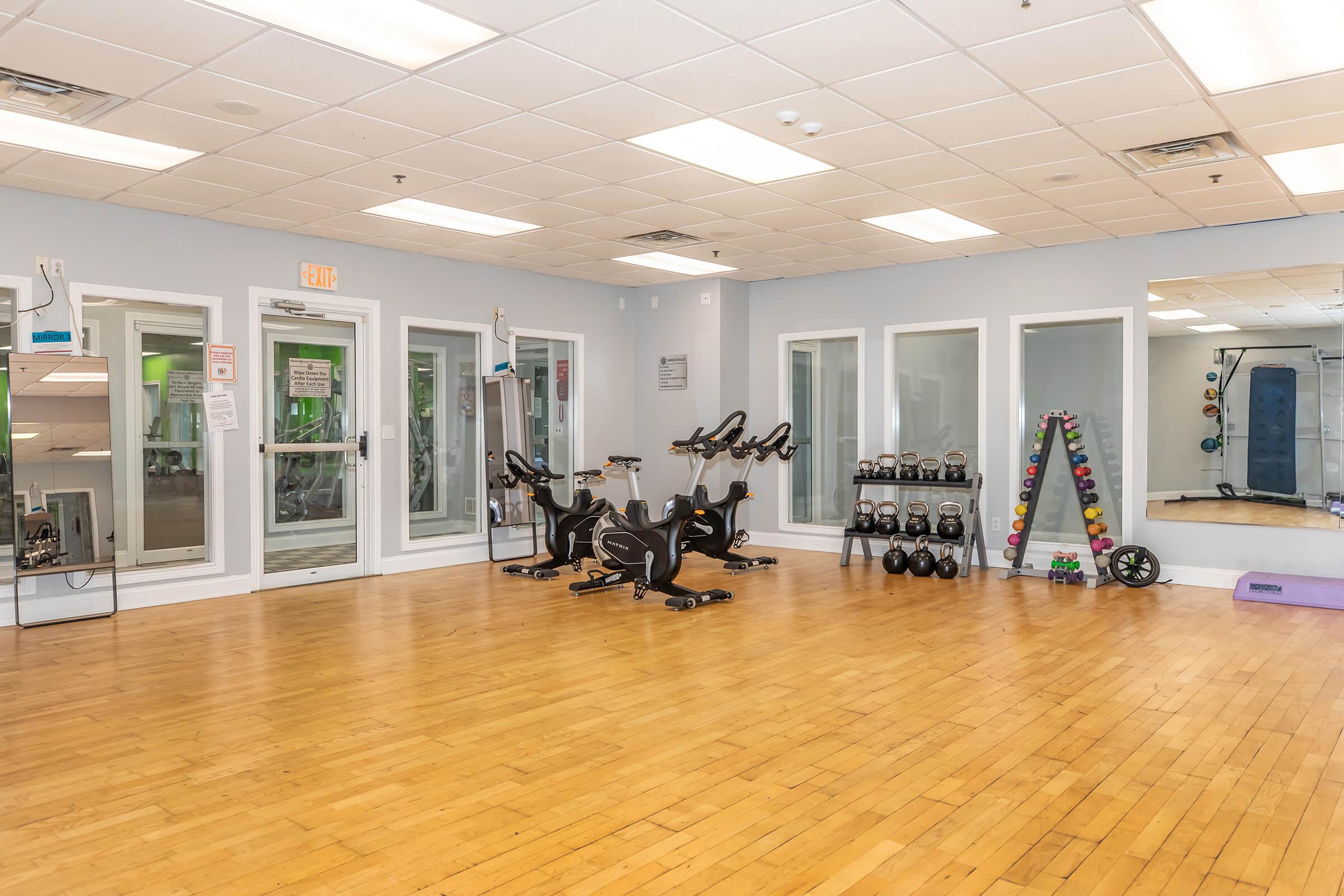 A bright and spacious fitness room featuring several stationary bicycles, a rack of dumbbells, kettlebells, and exercise mats. Large windows allow natural light to fill the space, creating an inviting atmosphere for workout activities.