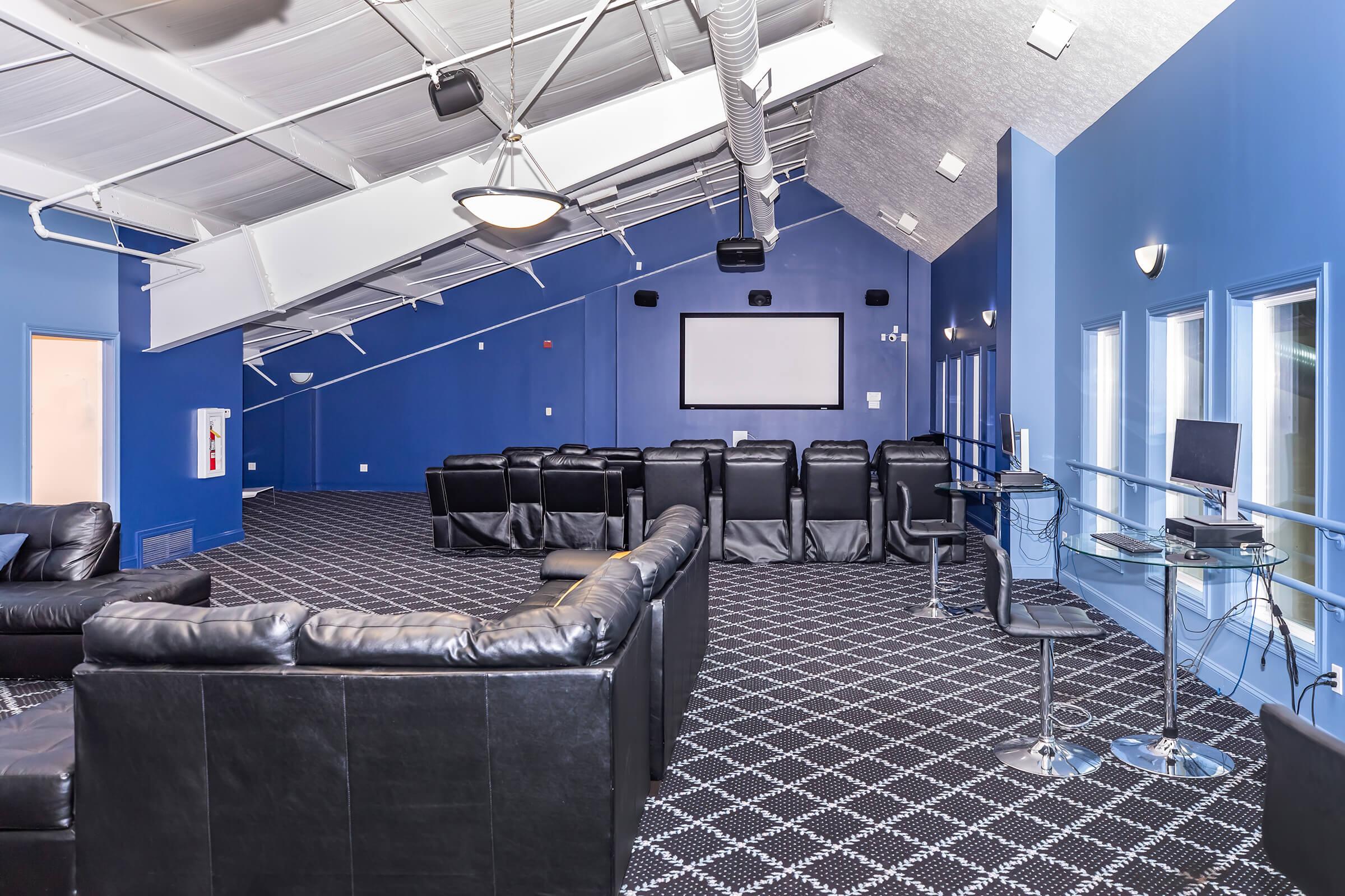 A spacious modern lounge area with blue walls and a high ceiling. The room features black leather seating arranged for viewing a large screen, with a checkered carpet. There are also glass-top tables with high stools and a small desk setup against one wall, creating a comfortable and inviting atmosphere.
