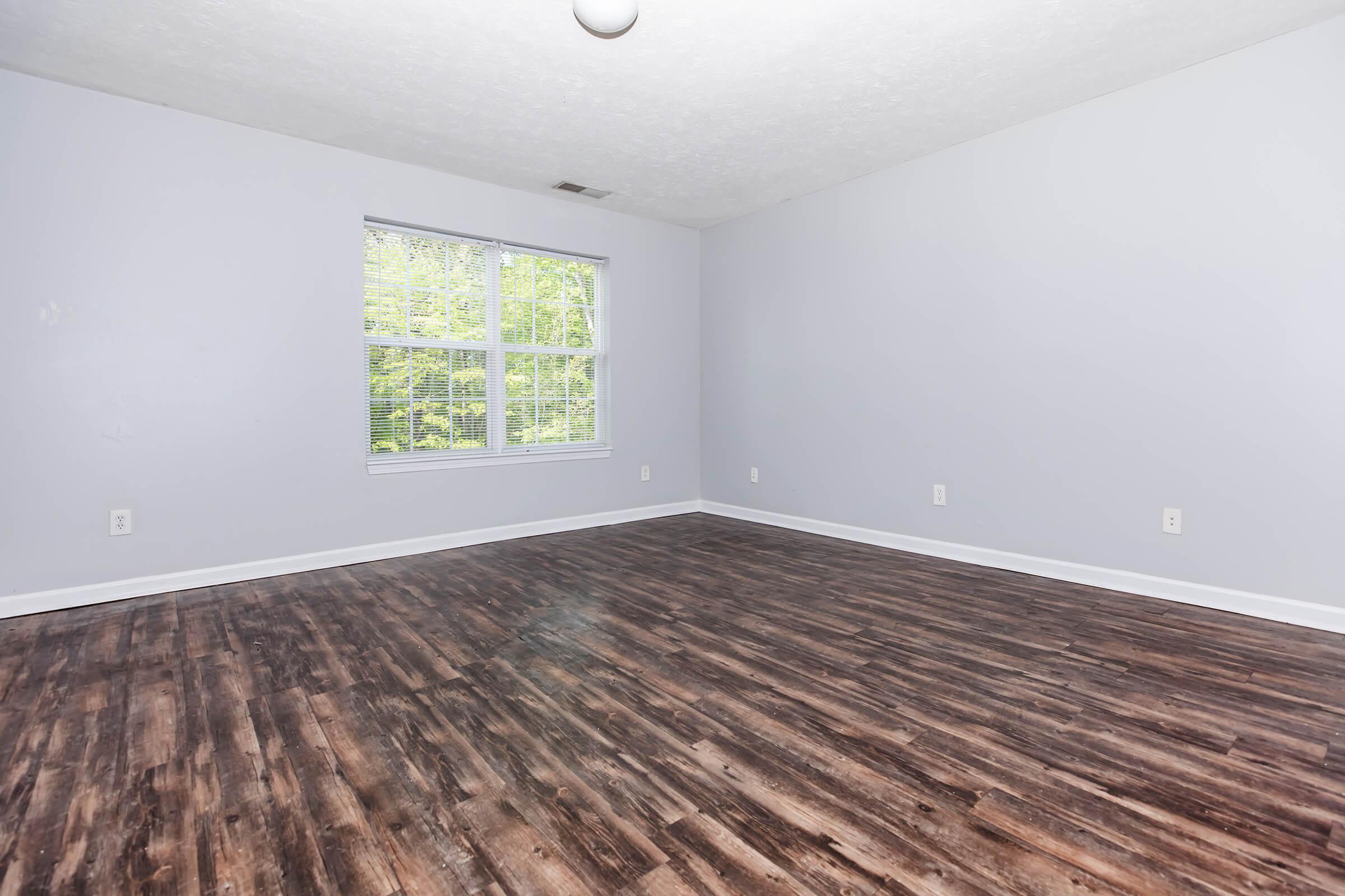 A spacious, empty room with light gray walls and a large window allowing natural light in. The floor features dark wood laminate, and the ceiling has a simple light fixture. The overall atmosphere is bright and open, suitable for various uses or decoration.