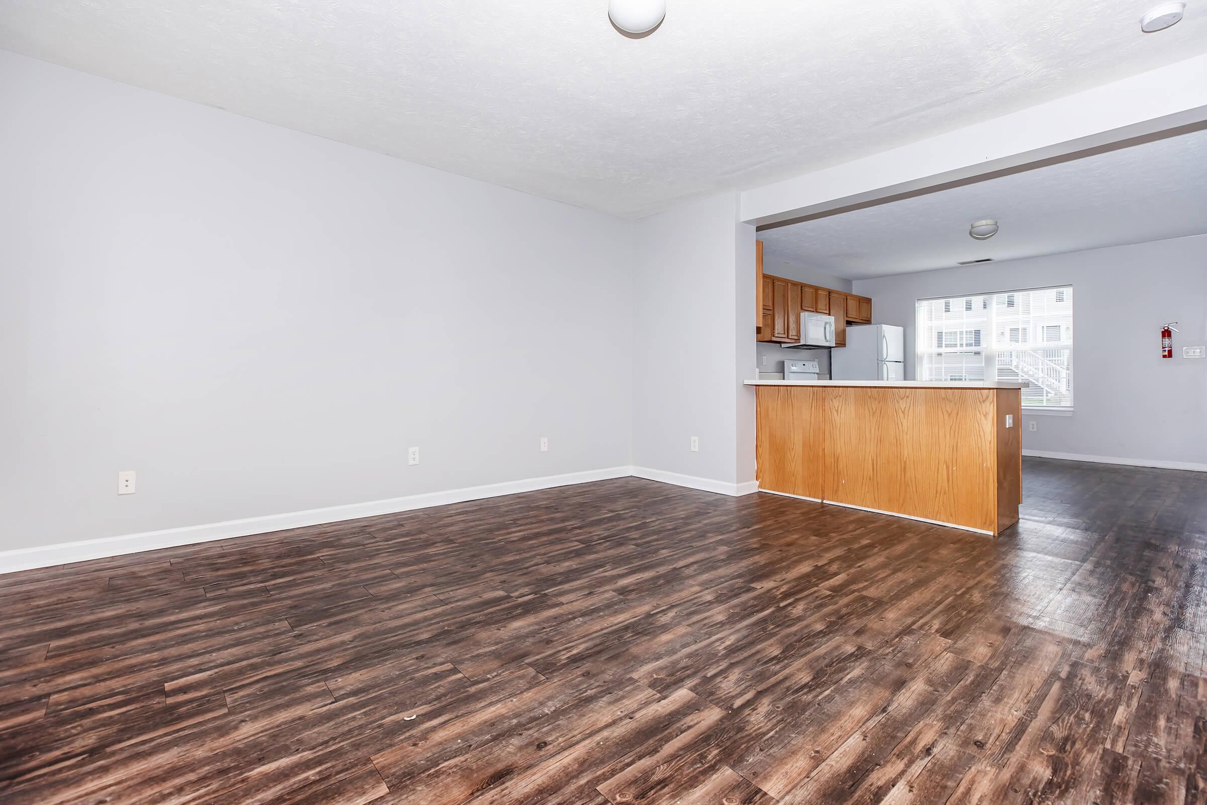 Spacious living area featuring hardwood-style flooring, light gray walls, and a kitchen bar counter that opens into the kitchen. The kitchen includes wooden cabinets and white appliances, creating a bright and inviting atmosphere. Natural light floods the space through large windows.