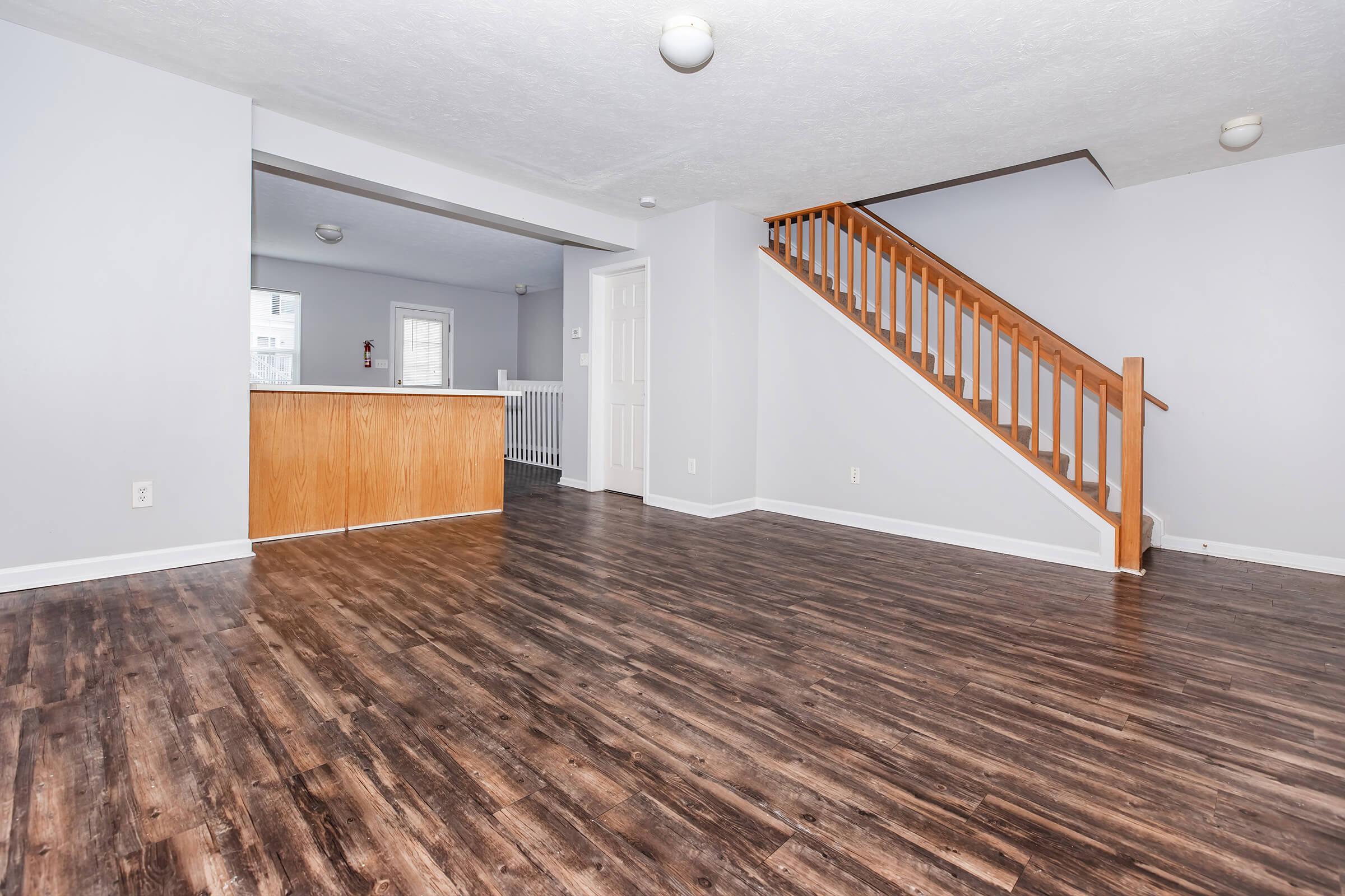 A spacious, well-lit room featuring hardwood floors, light gray walls, and a staircase leading to the upper level. In the background, an open kitchen area with wooden cabinetry is visible. The space is unfurnished, creating a clean and inviting atmosphere.