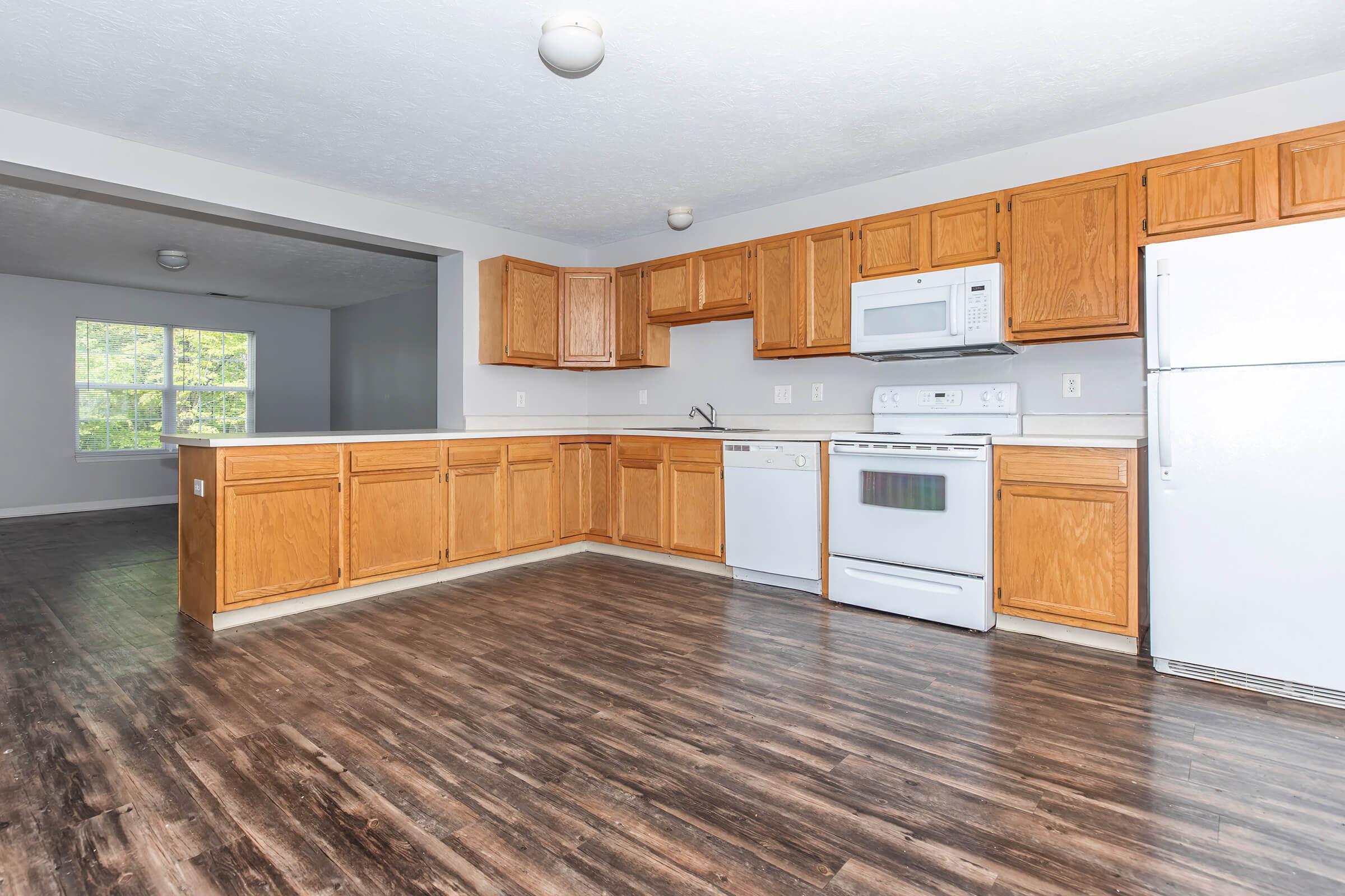 Spacious kitchen featuring wooden cabinets, white appliances, and a large window with natural light. Includes a refrigerator, microwave, dishwasher, and stove. The flooring is dark laminate, and an open layout connects to a living area.