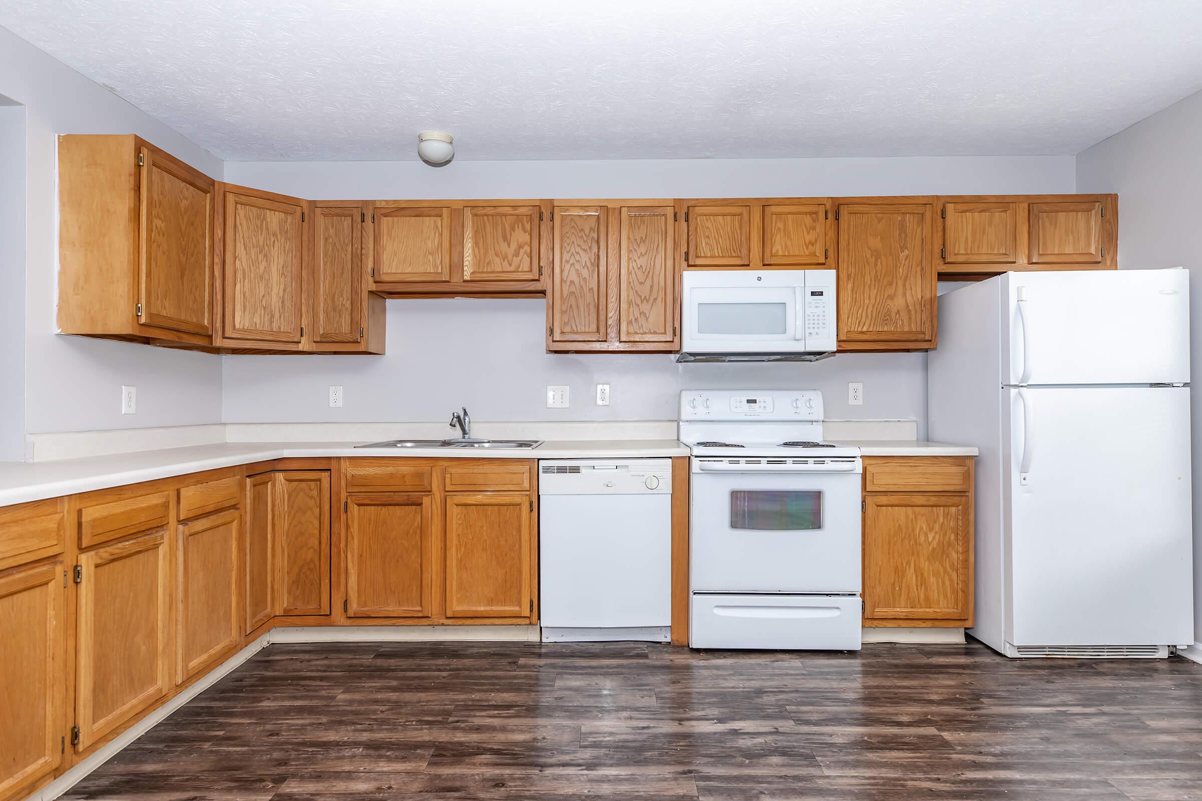 A modern kitchen featuring wooden cabinets, a white microwave, an oven, and a dishwasher. The countertops are light-colored, and a white refrigerator stands against one wall. The floor is made of dark wood, giving the space a cozy appearance. The walls are painted a soft gray.