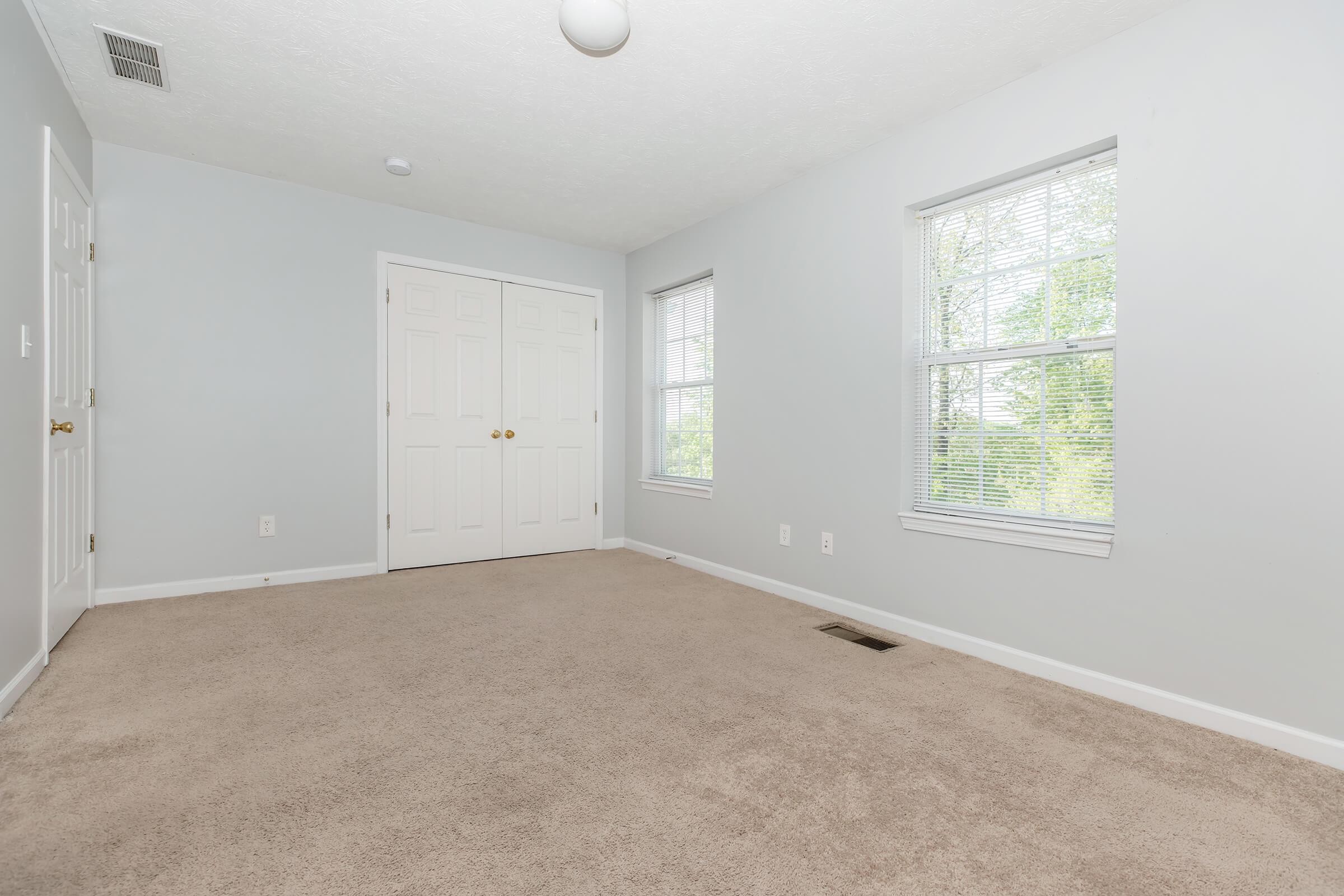 A bright, empty room with light gray walls and beige carpet. Two windows allow natural light to pour in, and a ceiling light fixture is visible. There are two closed doors on the left side, and a vent on the floor near the bottom right corner.