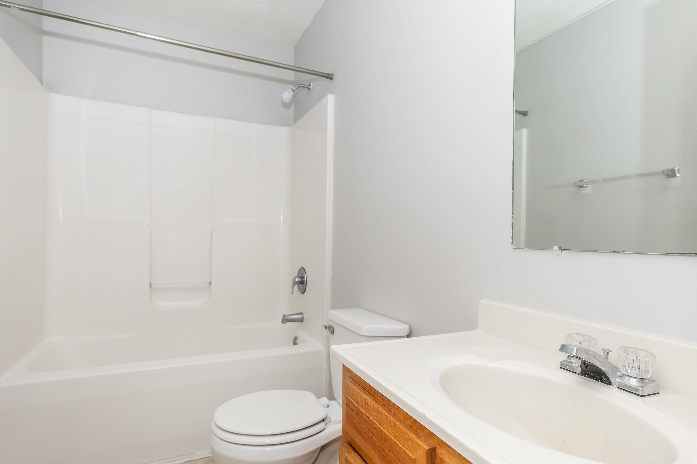 A clean, simple bathroom featuring a white tub and shower combination, a toilet, and a sink with a wooden cabinet underneath. The walls are painted light gray, and a mirror is mounted above the sink. Natural light illuminates the space, creating a fresh and airy atmosphere.