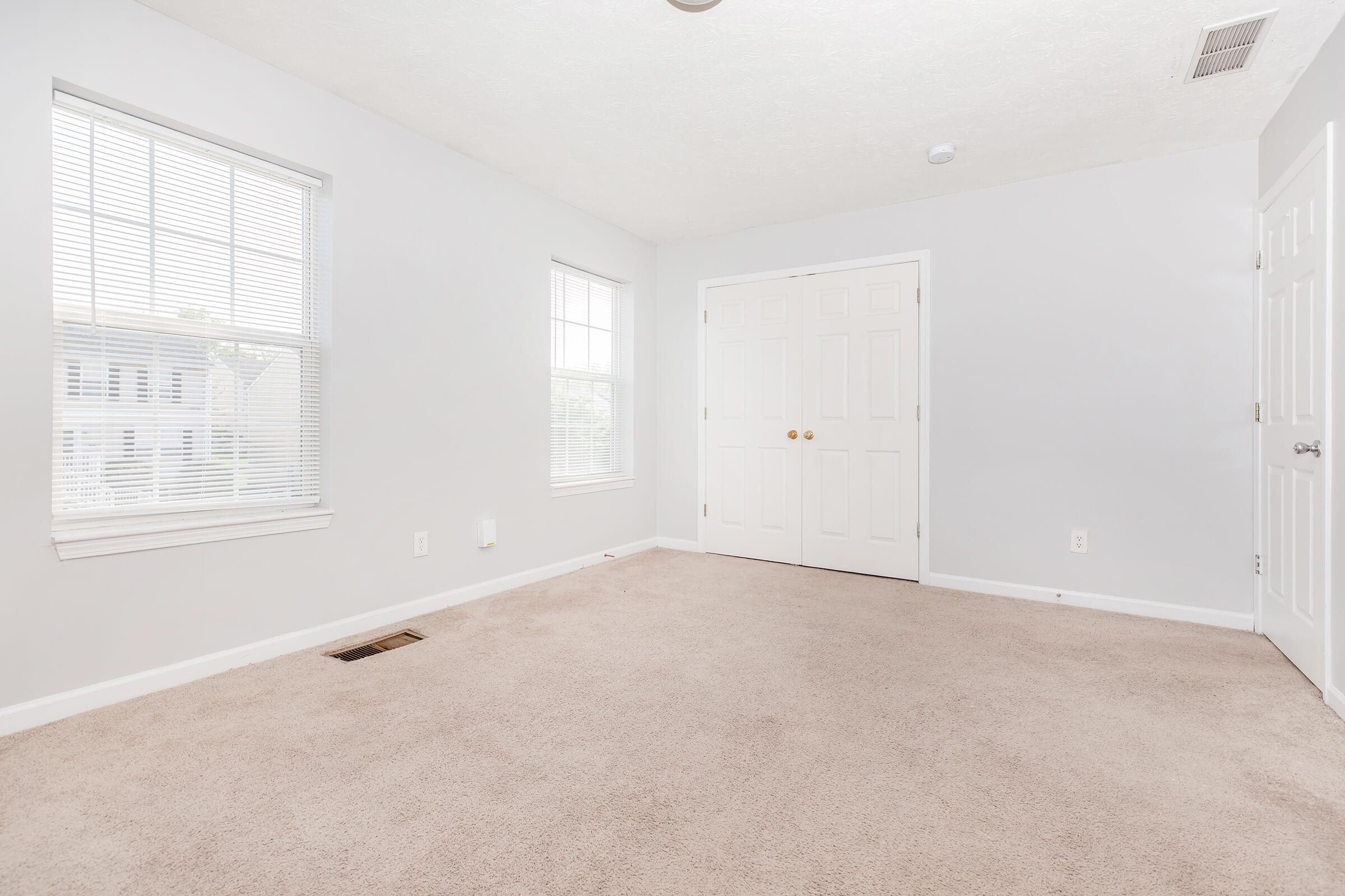 A spacious, empty room with light gray walls and soft beige carpet. There are two windows with blinds allowing natural light, and a double closet with white doors on one side. The room is well-lit and sparsely furnished, creating a clean and minimalist look.