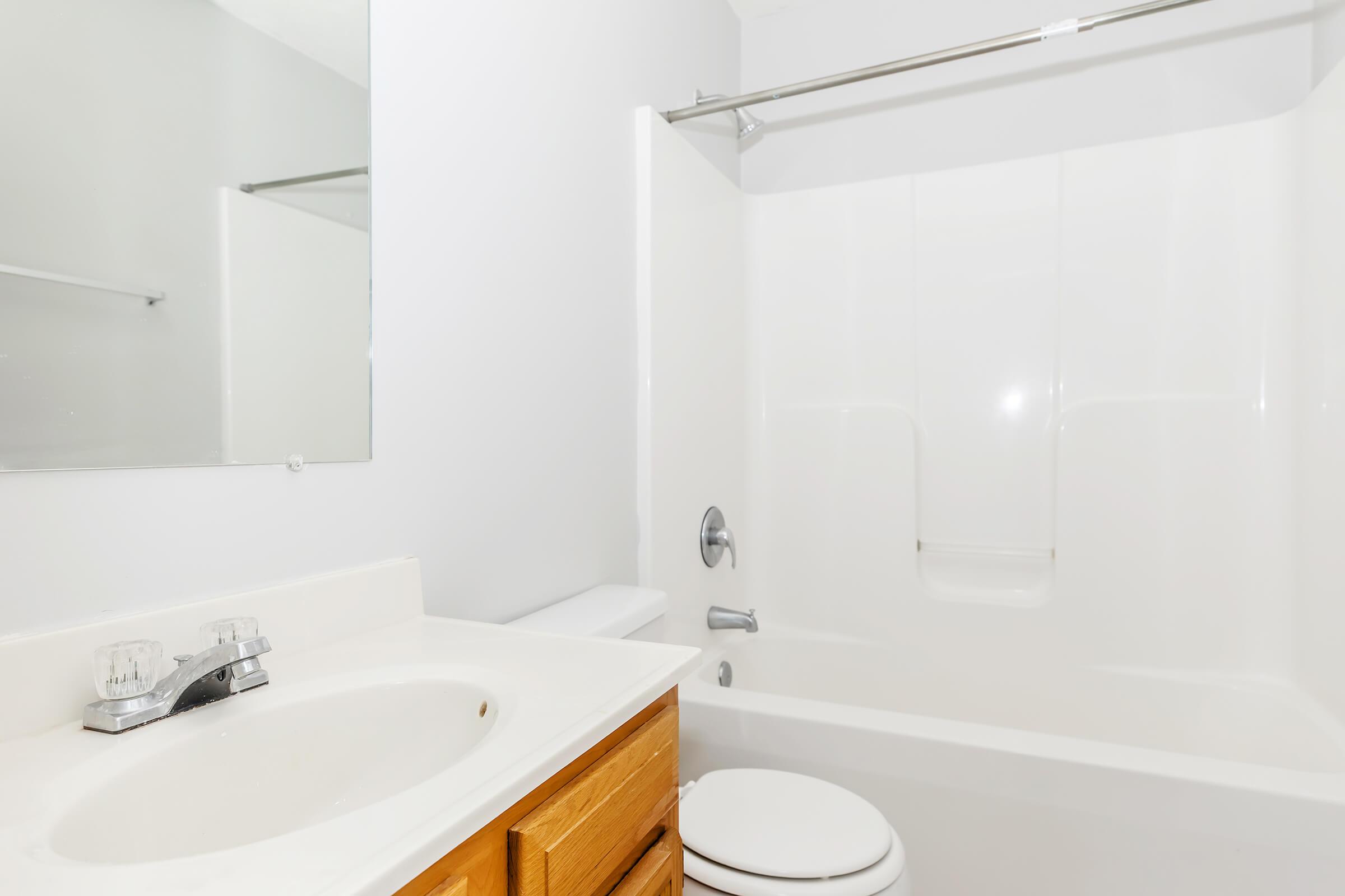 A clean, modern bathroom featuring a white bathtub and shower combination, a single sink with a faucet, and wooden cabinetry. A large mirror is mounted above the sink, and the walls are painted white, creating a bright and open space.