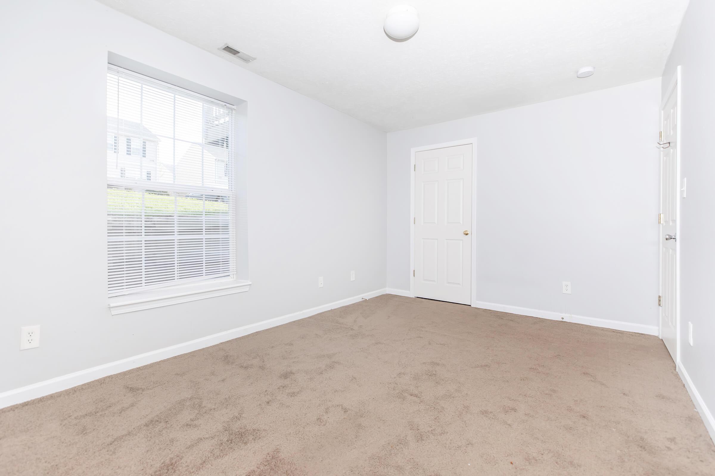 A vacant room with light gray walls and beige carpet. There is a window on one wall allowing natural light, and a closed white door on another wall. The room is spacious and uncluttered, creating a minimalist atmosphere. A ceiling light fixture is visible, and the overall setting appears well-maintained.
