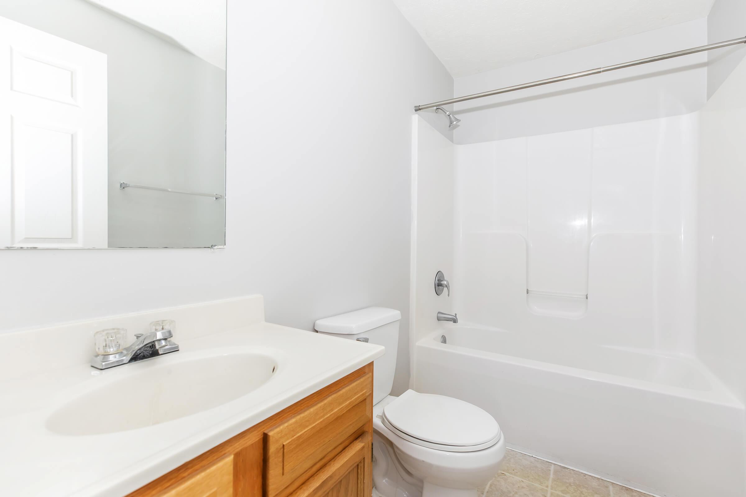 A clean and simple bathroom featuring a white bathtub with a shower, a toilet, and a wooden vanity with a sink. The walls are painted light gray, and there is a large mirror above the sink. The floor has light-colored tiles, contributing to a bright and airy atmosphere.