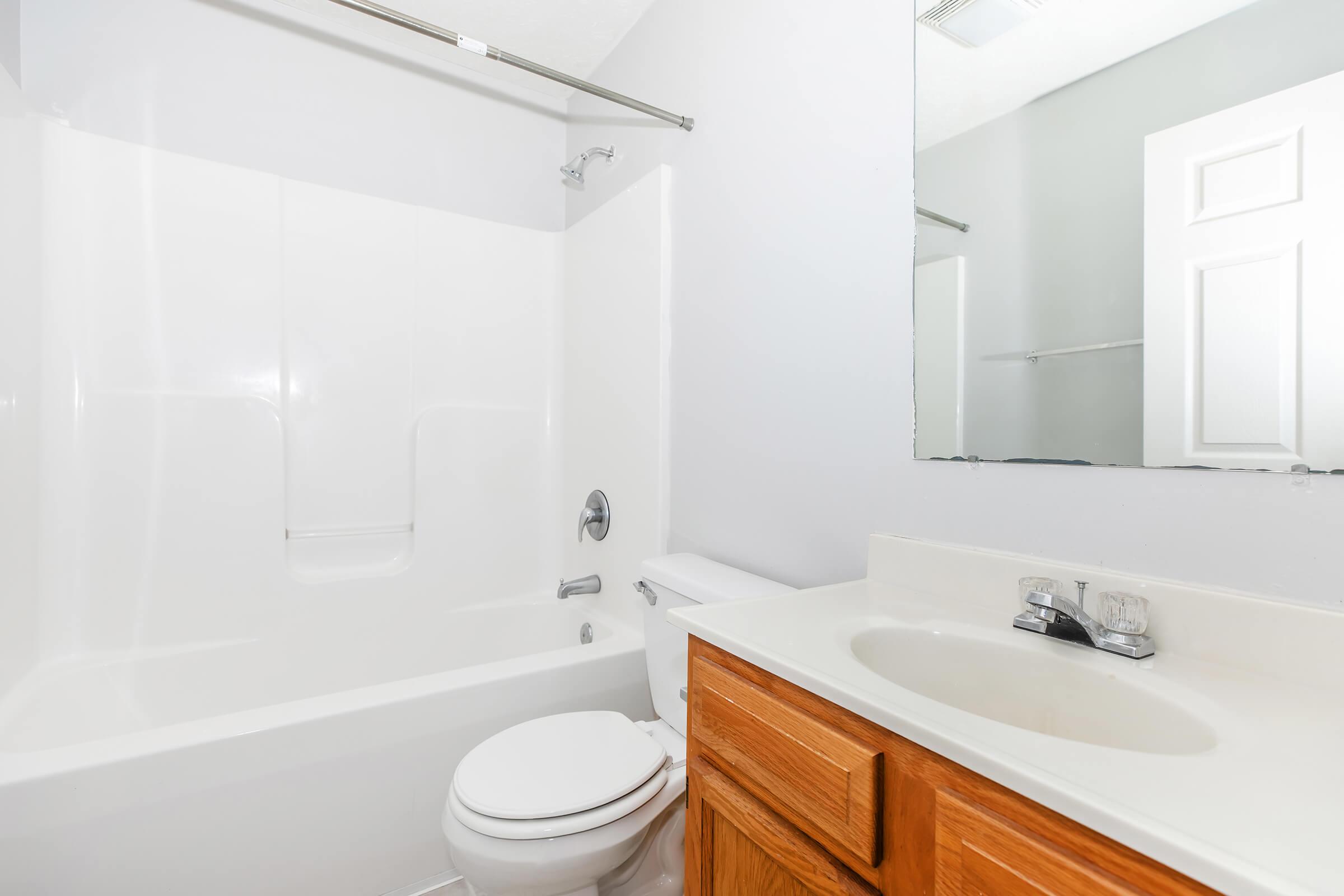 A clean and modern bathroom featuring a white tub and shower combination with a silver showerhead, a toilet, and a light-colored countertop with a sink. The walls are painted in a neutral color, and there's a large mirror above the sink with a white door visible in the background.