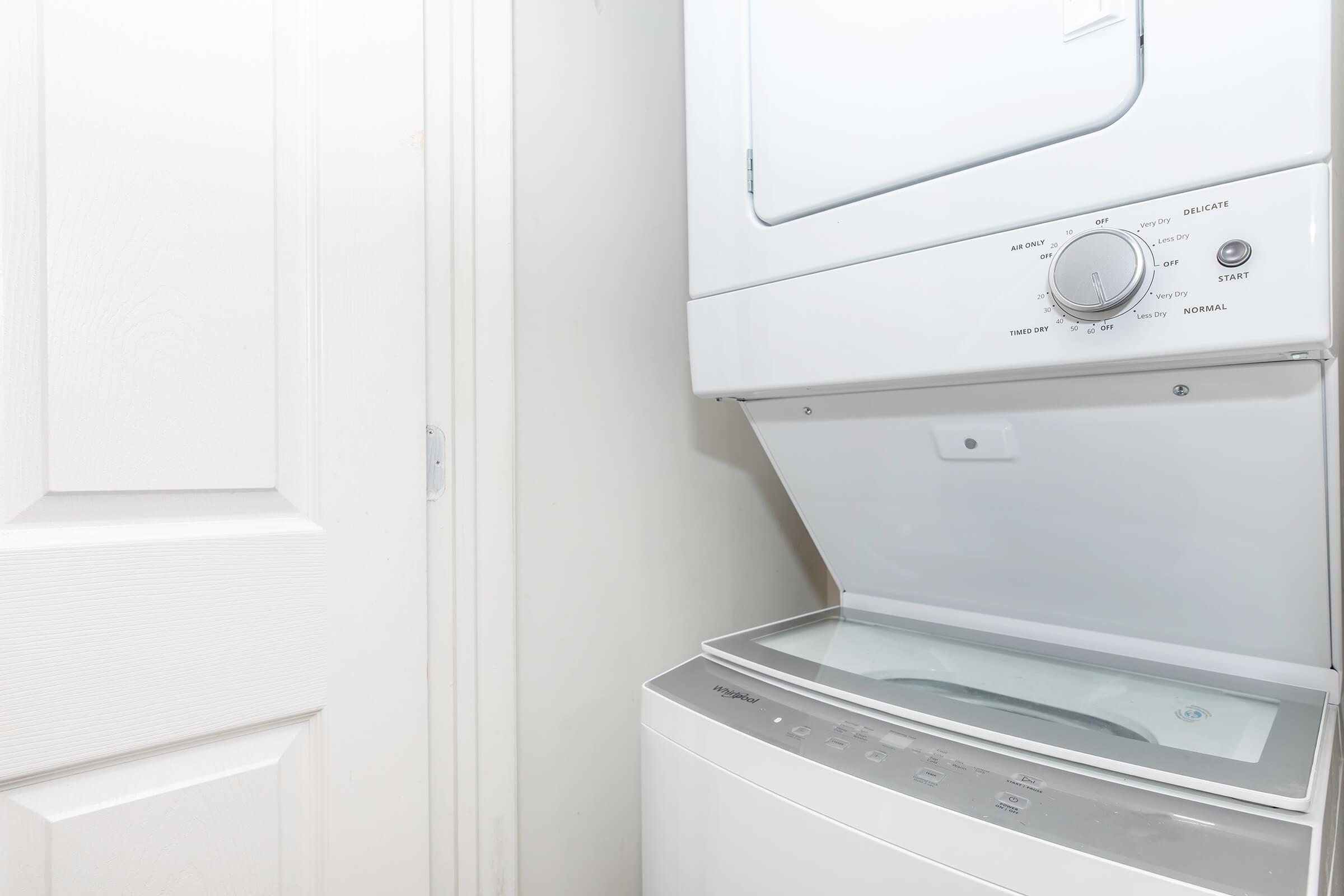 A stacked washer and dryer unit in a laundry room, with the dryer on top and the washer below. The dryer is slightly open, revealing the drum inside. A door is visible in the background, suggesting the laundry area is located in a small space. The walls are light-colored, giving a clean and modern appearance.