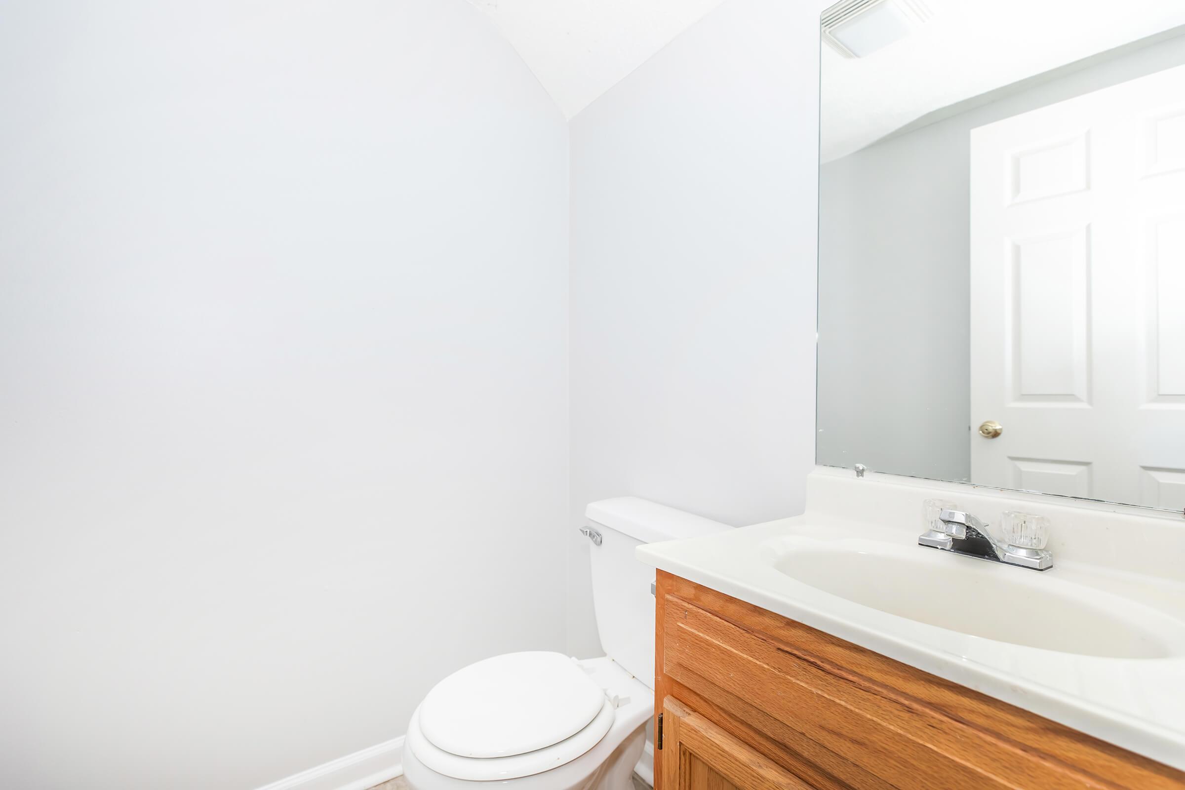 A clean and minimalistic bathroom featuring a white toilet, a wooden vanity with a sink, and a large mirror. The walls are painted in a light gray color, and there is a door leading to another room in the background. The space appears well-lit and tidy.