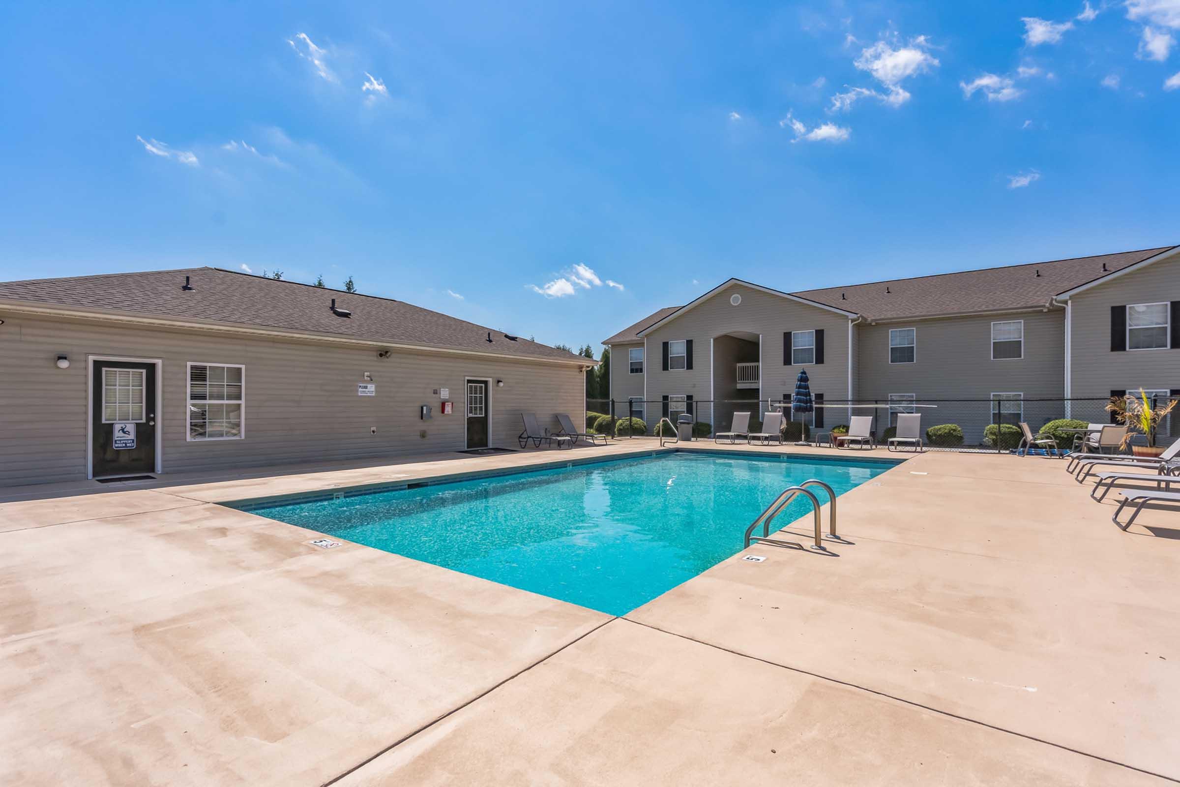 Outdoor pool area at an apartment complex, featuring a clear blue pool surrounded by a concrete deck. There are lounge chairs and tables nearby, with two adjacent buildings in the background under a bright blue sky.