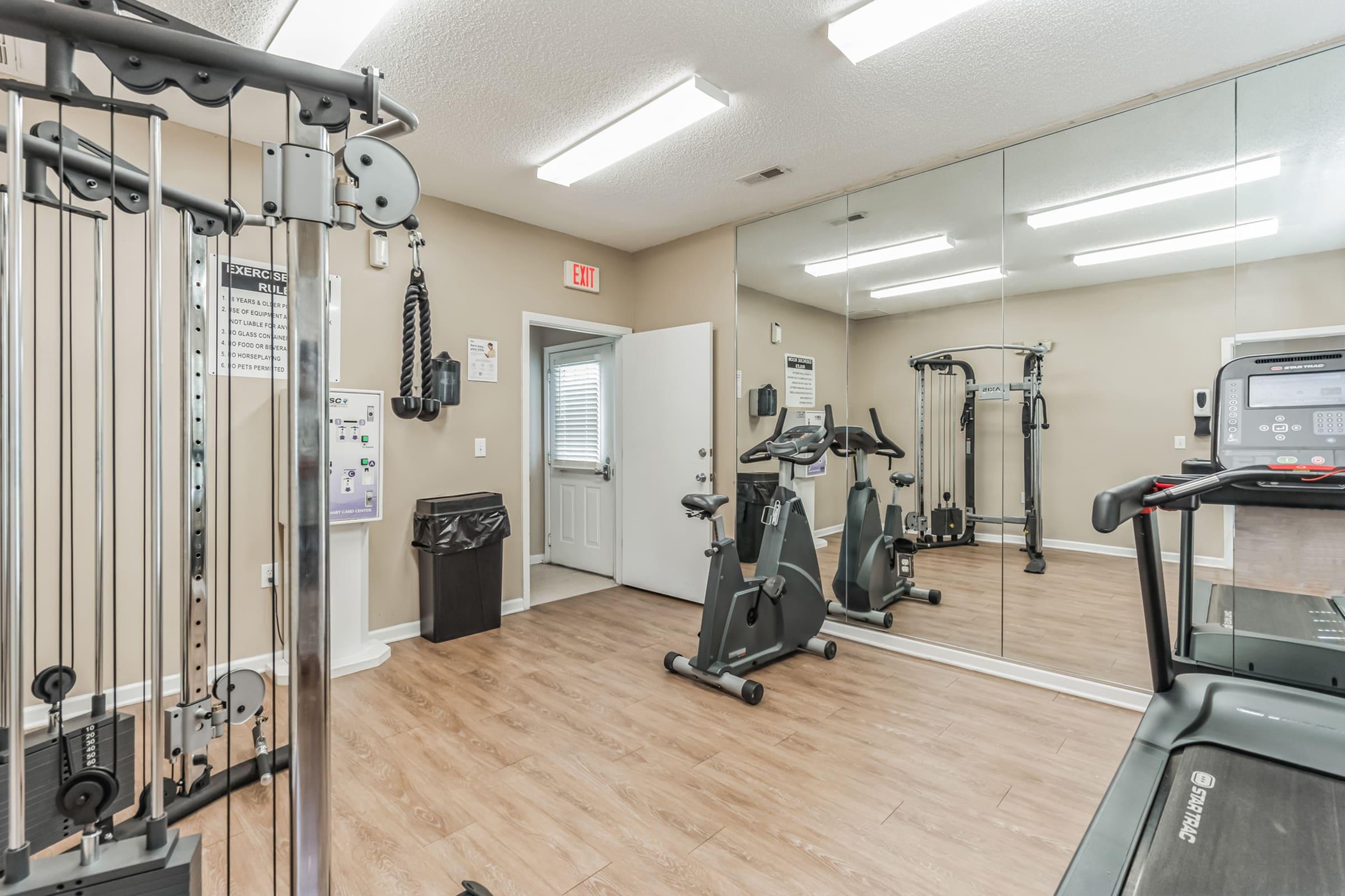 A fitness room featuring various exercise equipment, including a treadmill, stationary bikes, and a weight machine. The space is well-lit with mirrors on one wall and a door leading outside. There is also a trash can and fitness instructions on the wall. The flooring appears to be wood.