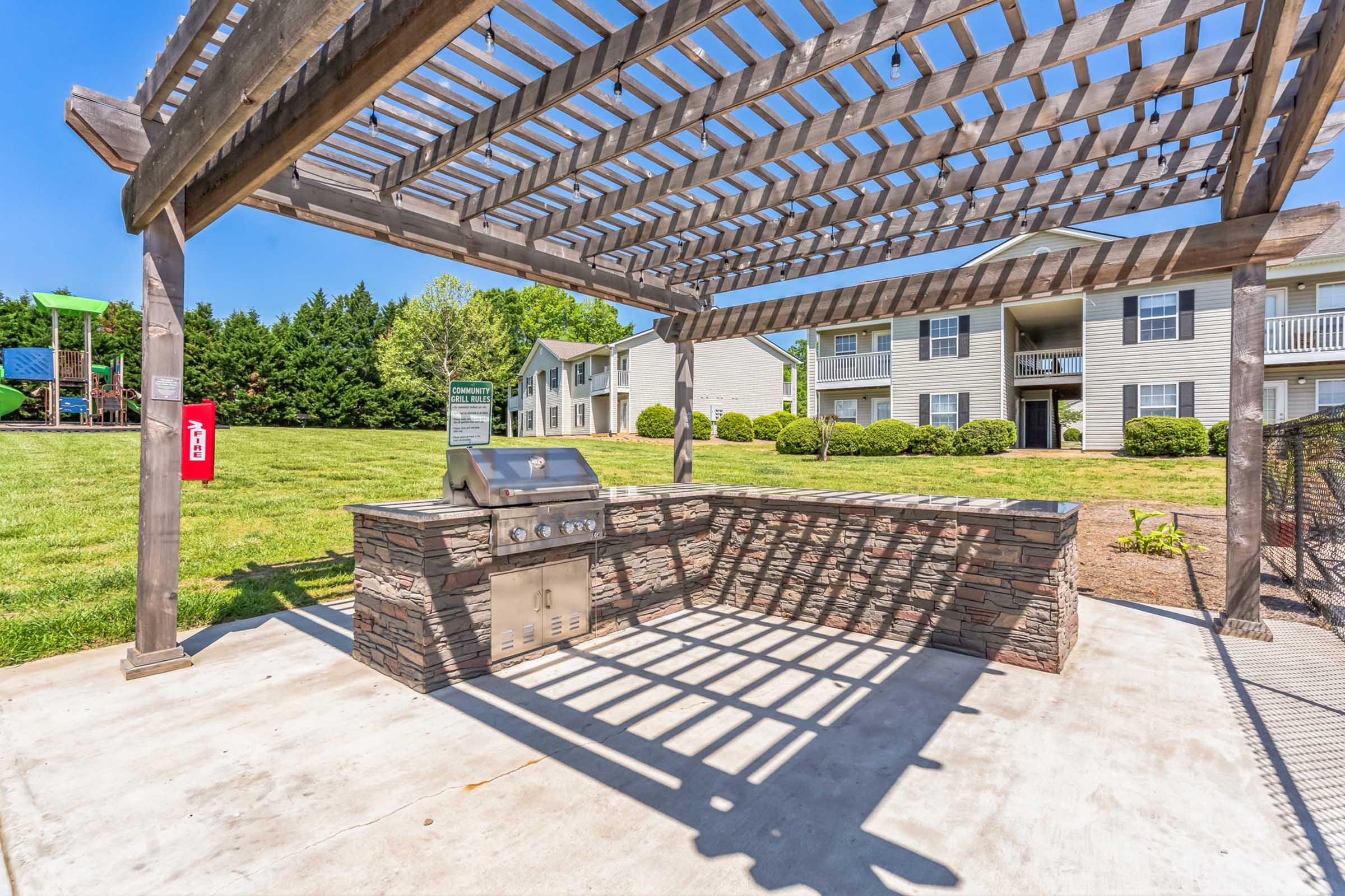 A shaded outdoor grilling area featuring a stone barbecue grill beneath a wooden pergola. In the background, there are well-maintained grassy areas and several apartment buildings. The scene is bright and inviting, perfect for outdoor cooking and gatherings.