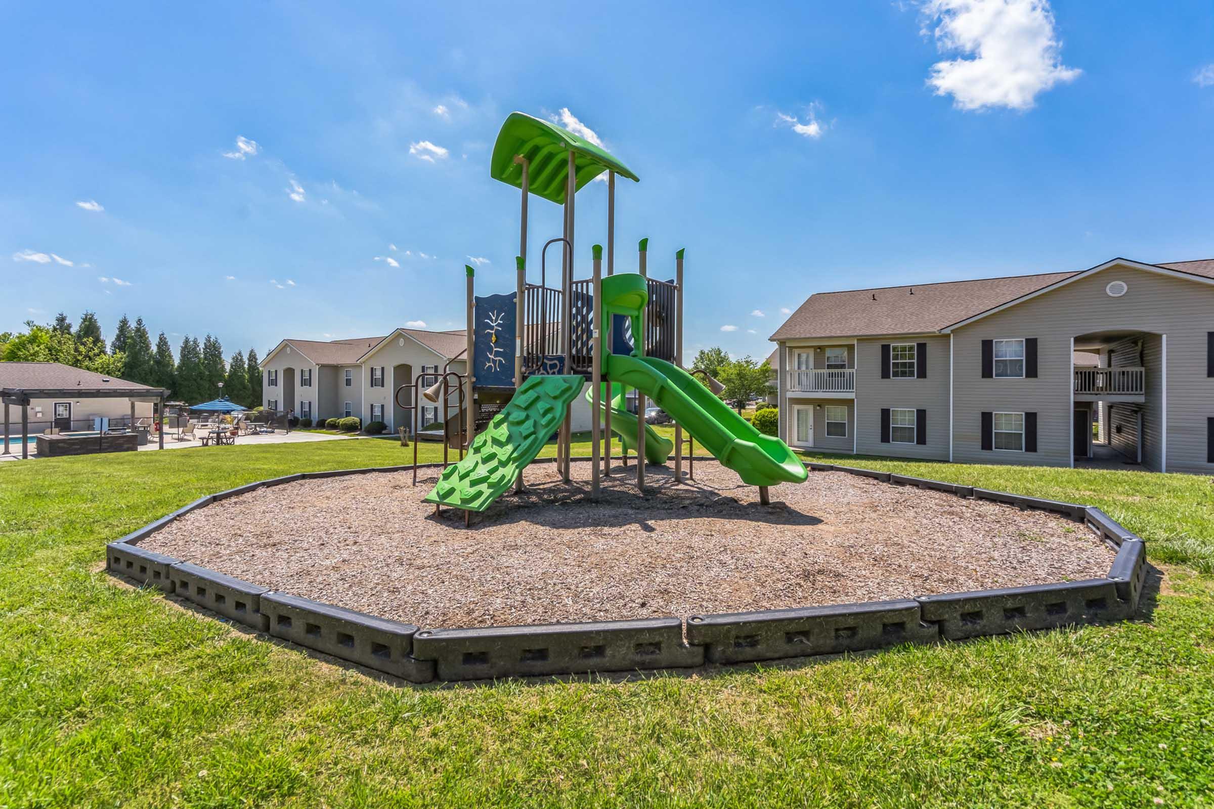 A colorful playground featuring a green slide and climbing structure, set on a bed of gravel. The play area is surrounded by grass and is adjacent to residential buildings with balconies under a clear blue sky.