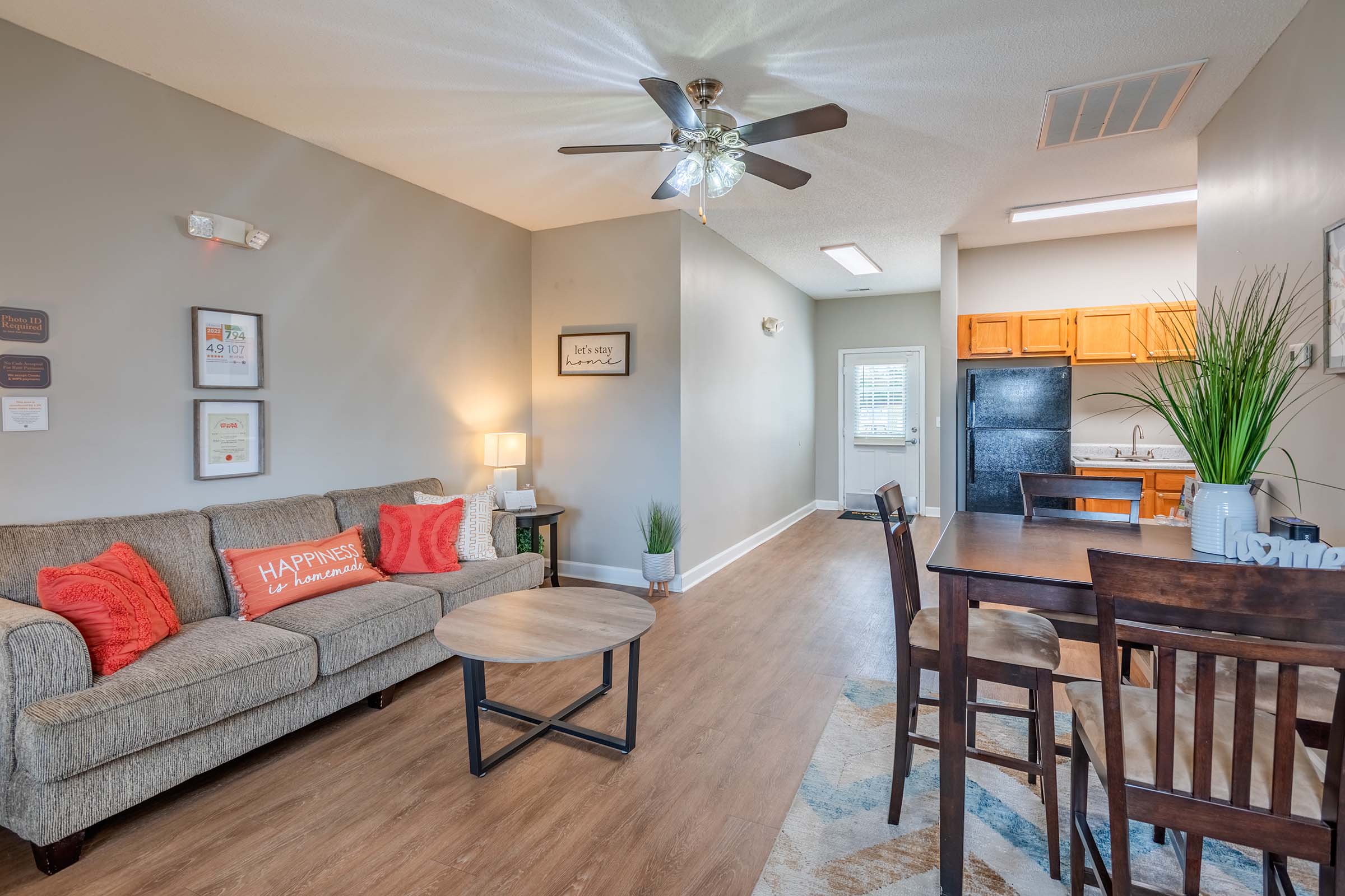 A cozy living room featuring a beige sofa with colorful cushions, a round coffee table, and a dining area with wooden chairs. The space is bright and welcoming, with a ceiling fan and modern kitchen visible in the background, creating a warm, inviting atmosphere.