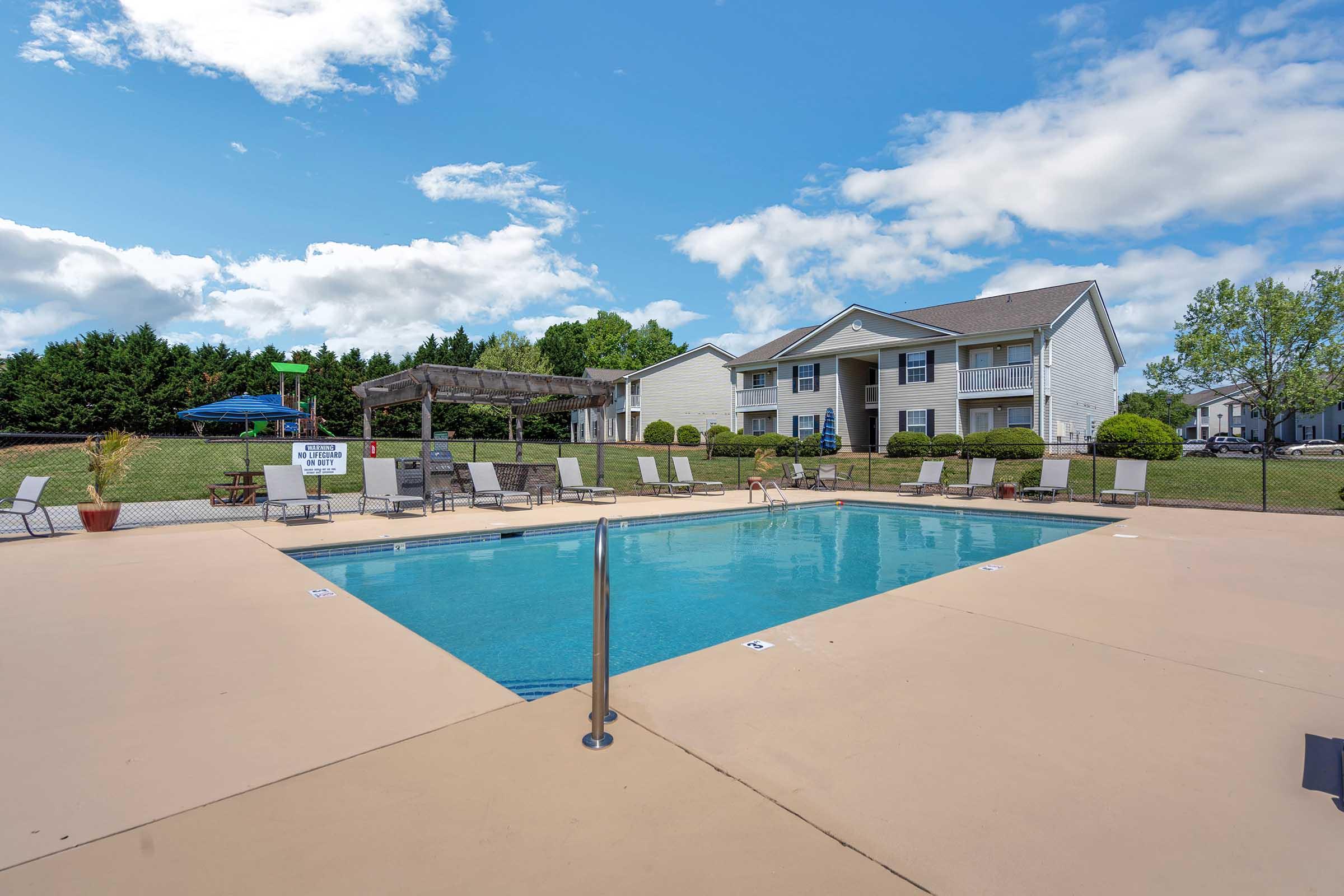 A clear blue swimming pool surrounded by lounge chairs and a patio area. In the background, there are residential buildings and green trees under a partly cloudy sky. A gazebo can be seen near the pool, creating a relaxing outdoor atmosphere.