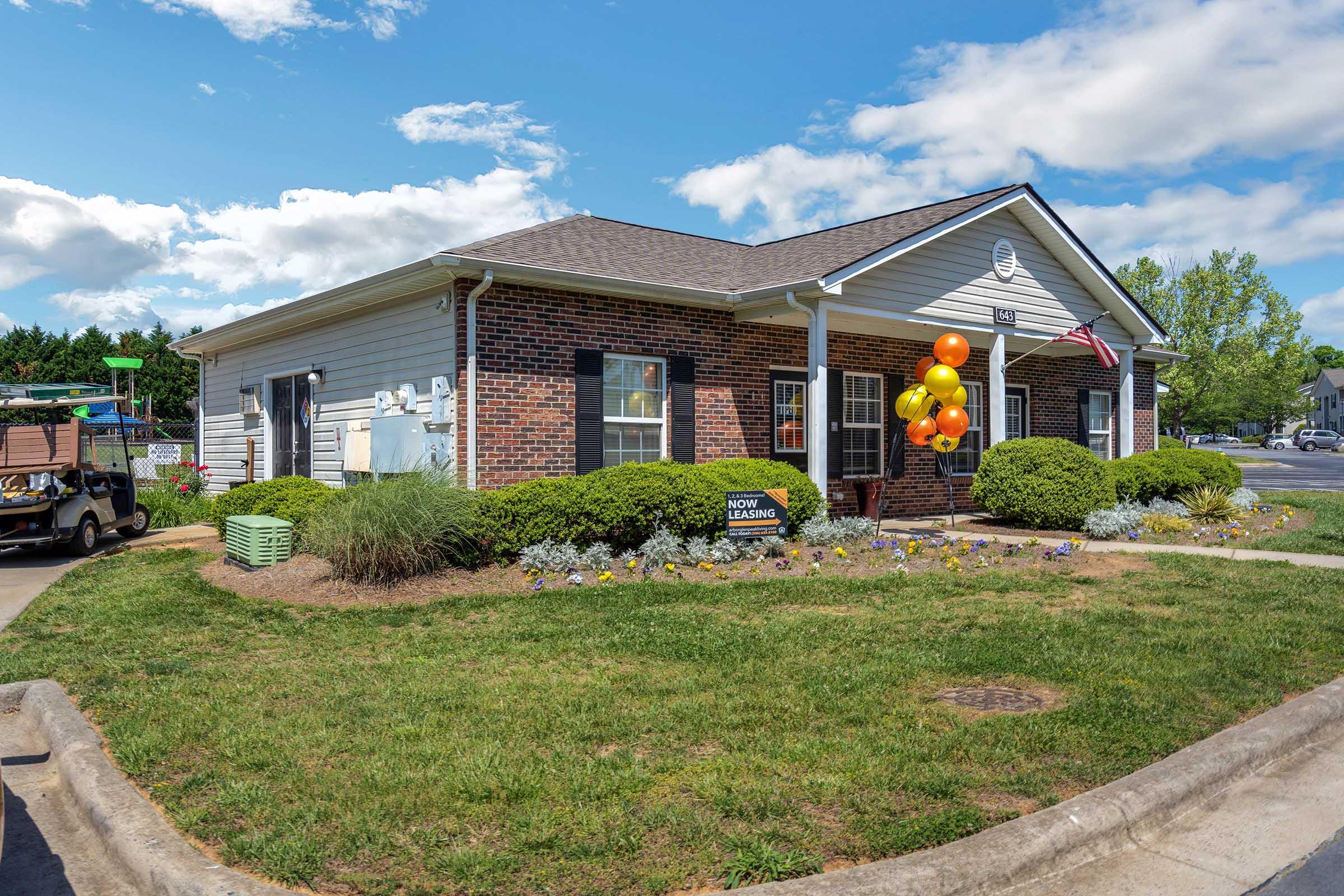 A single-story brick building with a gray wooden section, featuring a well-maintained lawn with colorful flowers and shrubs. Balloons in orange and yellow are tied to the entrance, which has a sign reading "GROW LEARNING." The sky is partly cloudy, and there are trees in the background, along with a golf cart parked nearby.