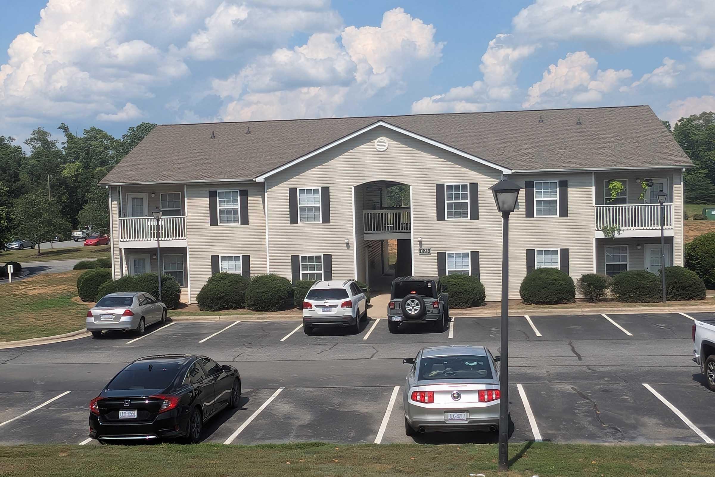 Two-story apartment building with beige siding and a gray roof, flanked by manicured bushes. Several parked cars in a lot in the foreground, including a black car, a gray sedan, and a white SUV. The sky is partly cloudy, and there are trees in the background.