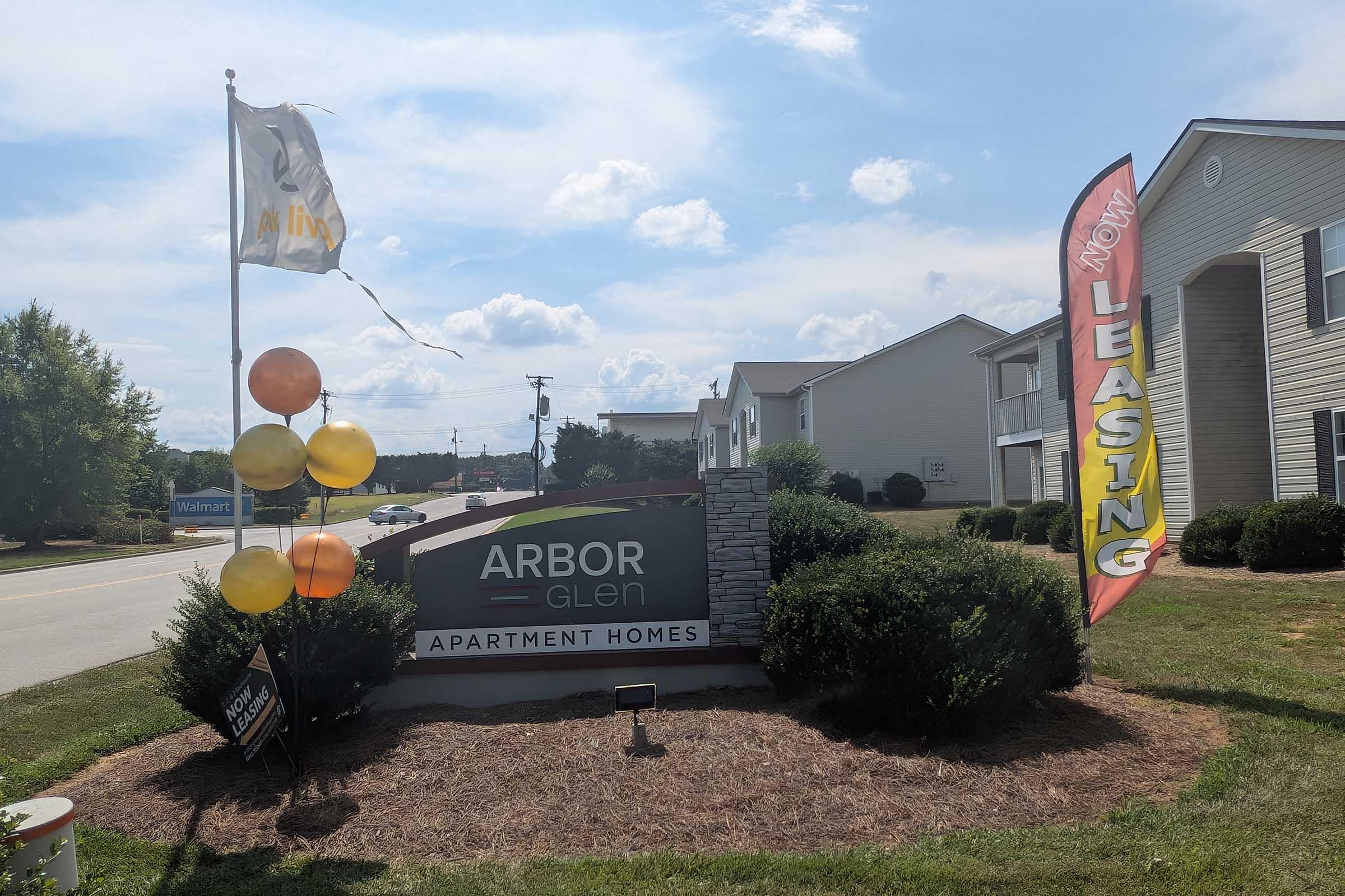 Sign for Arbor Glen Apartment Homes featuring a flag and a leasing banner. The entrance has decorative balloons and is surrounded by greenery, with a view of nearby buildings and a road. The sky is bright and partly cloudy, indicating a sunny day.