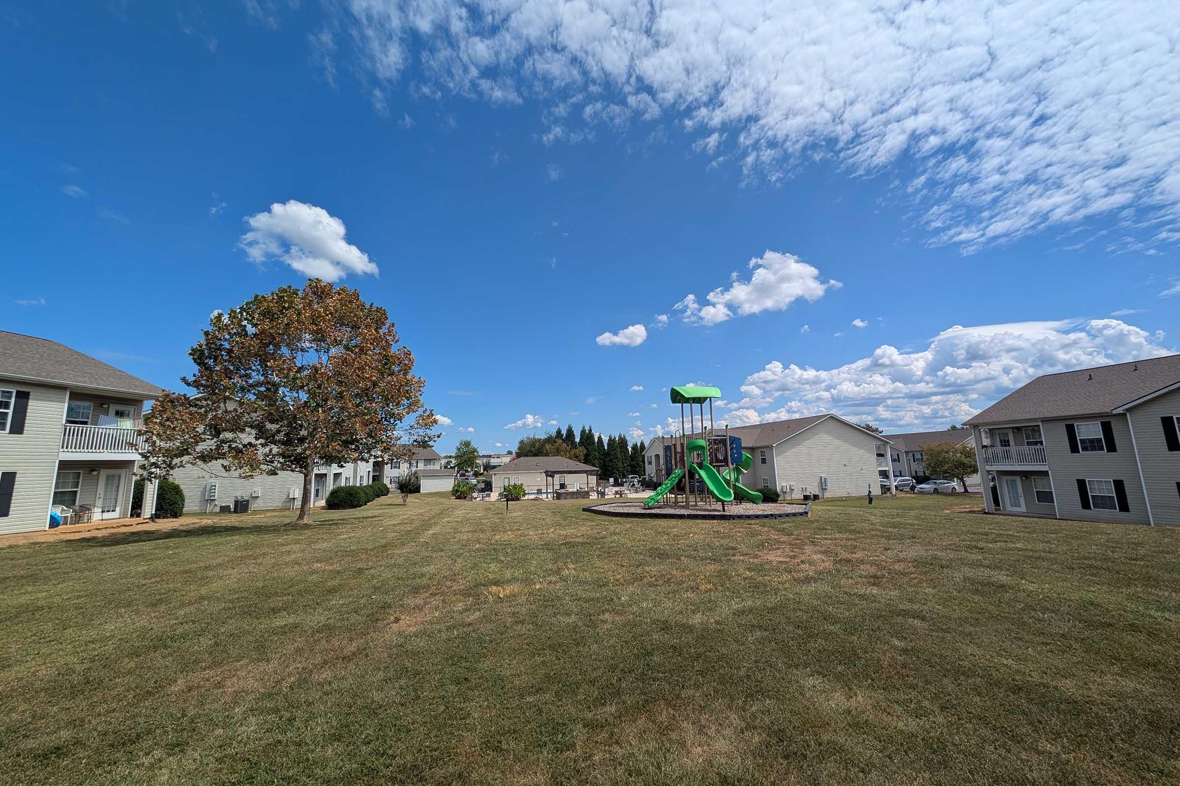 A grassy area surrounded by residential buildings under a clear blue sky with scattered clouds. In the center, there's a playground featuring a green slide and swings, with a large tree nearby that has autumn-colored leaves.