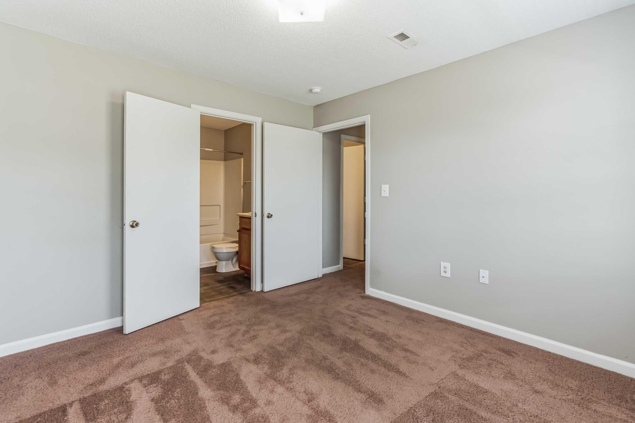 A spacious room with light brown carpet, featuring two open doors leading to adjacent spaces. One door opens to a bathroom with a toilet visible. The walls are painted in a neutral gray color, and there is a ceiling light fixture. Natural light may be entering from a window.