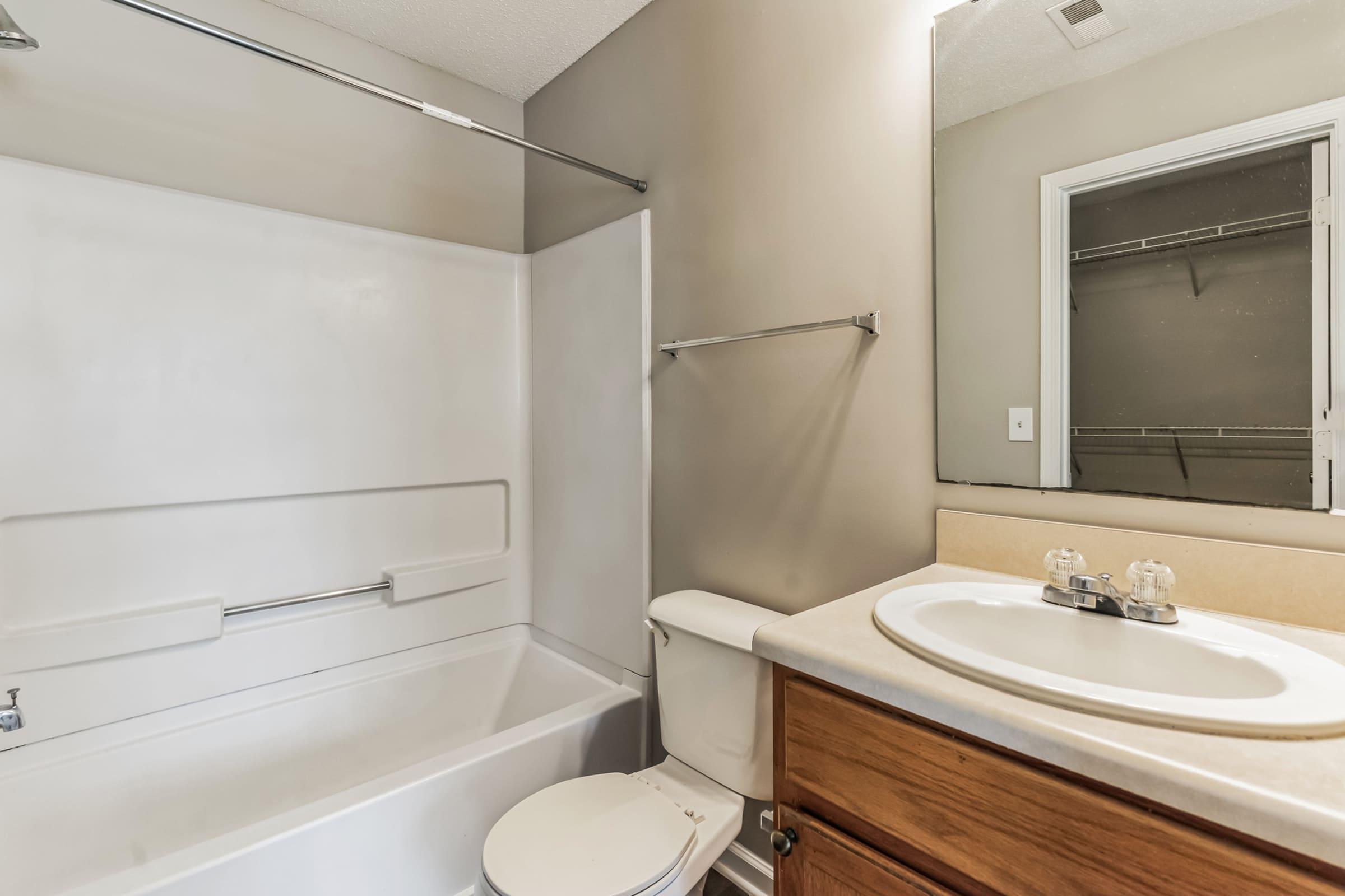 A clean bathroom featuring a white shower and bathtub combination, a toilet, and a sink with wooden cabinetry. The walls are painted in neutral tones, and there is a mirror above the sink. A small window can be seen in the background, providing natural light.