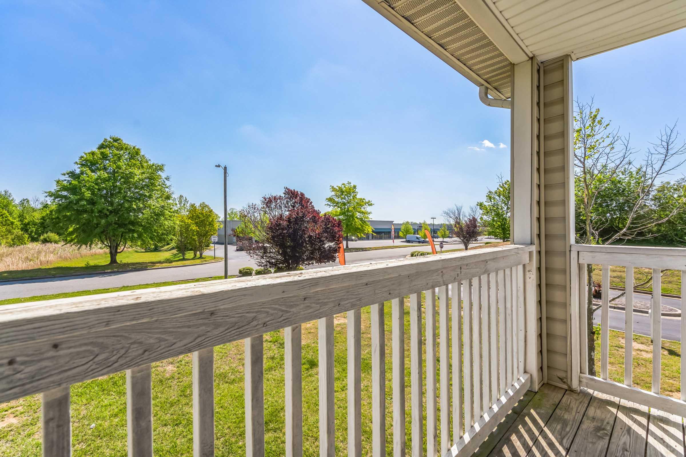 A view from a balcony overlooking a grassy area and trees, with a clear blue sky above. The scene includes a road in the distance and a few scattered buildings, creating a peaceful residential atmosphere. The balcony railing is wooden and highlights the outdoor setting.