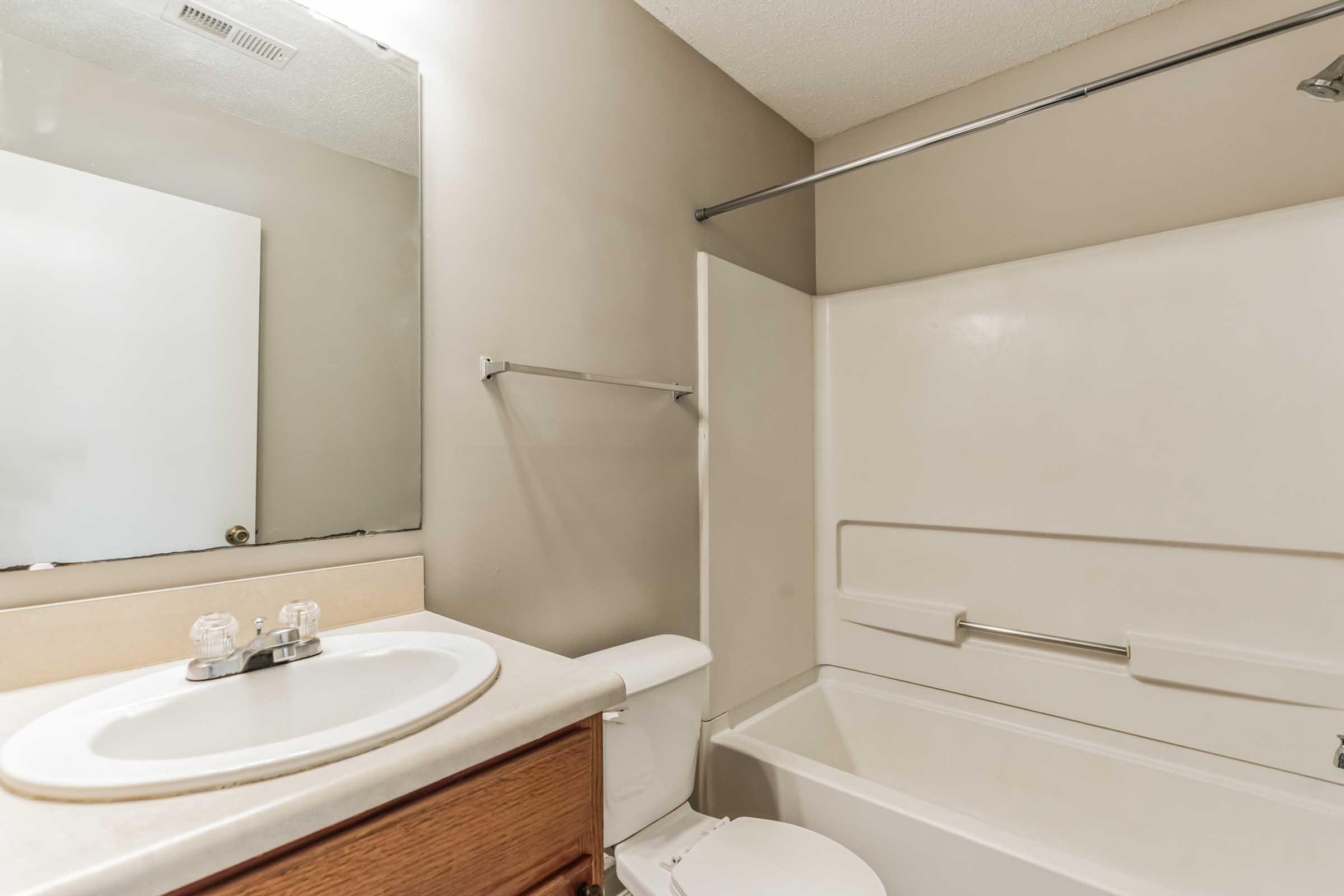 A clean and neutral bathroom featuring a white bathtub, a toilet, and a sink with a mirror above it. The walls are painted in a light beige color, and there is a shower curtain rod without a curtain. The overall design is simple and functional.