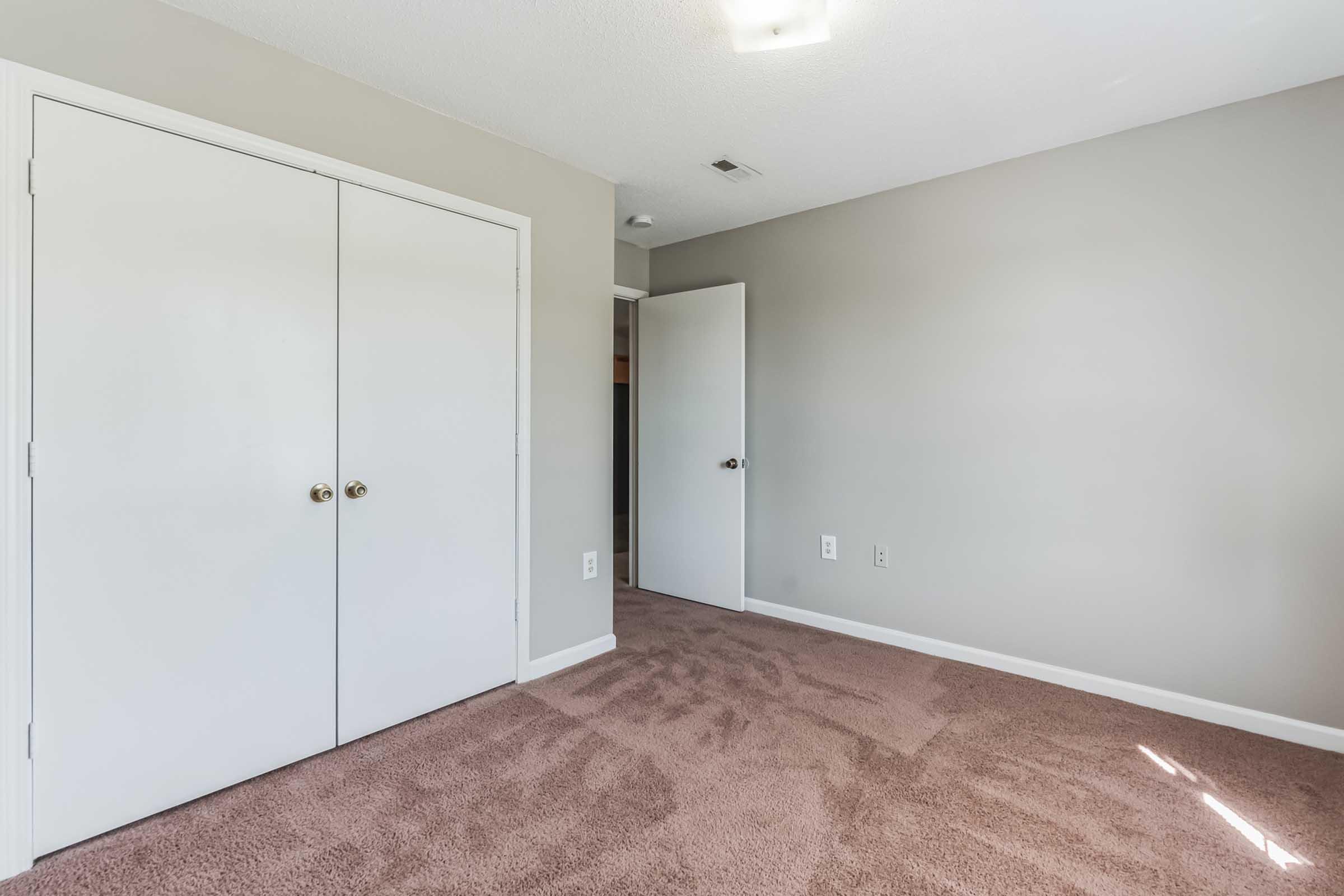 Empty room with beige carpet, light gray walls, and a closet with double white doors. A single door leads to another area. Natural light spills in from a nearby window, creating a bright and open atmosphere.