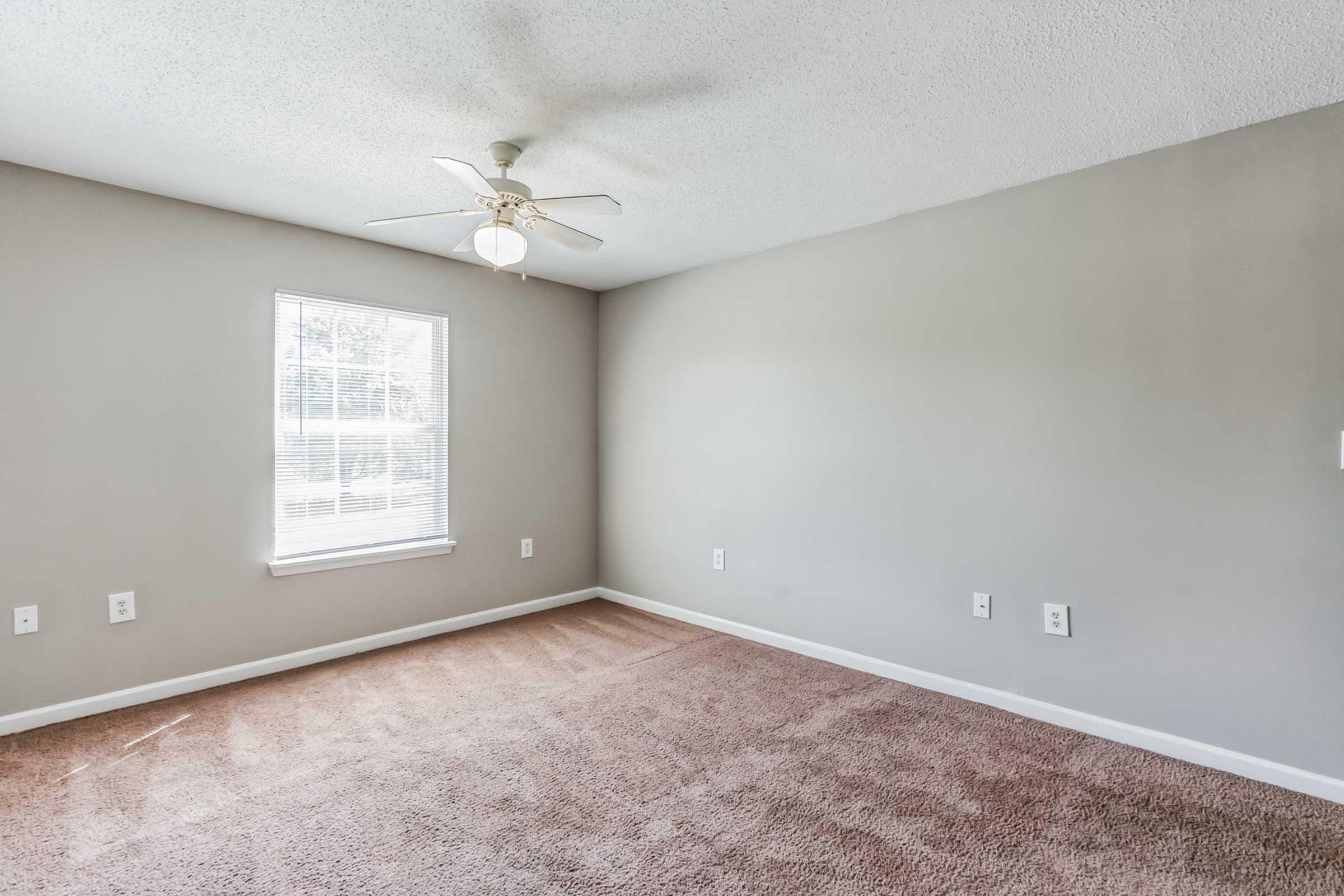 A spacious, unfurnished room with light beige carpet and light gray walls. A ceiling fan is mounted in the center, and there is a window on one wall allowing natural light to enter. The room features several electrical outlets along the walls, creating a bright and airy atmosphere.