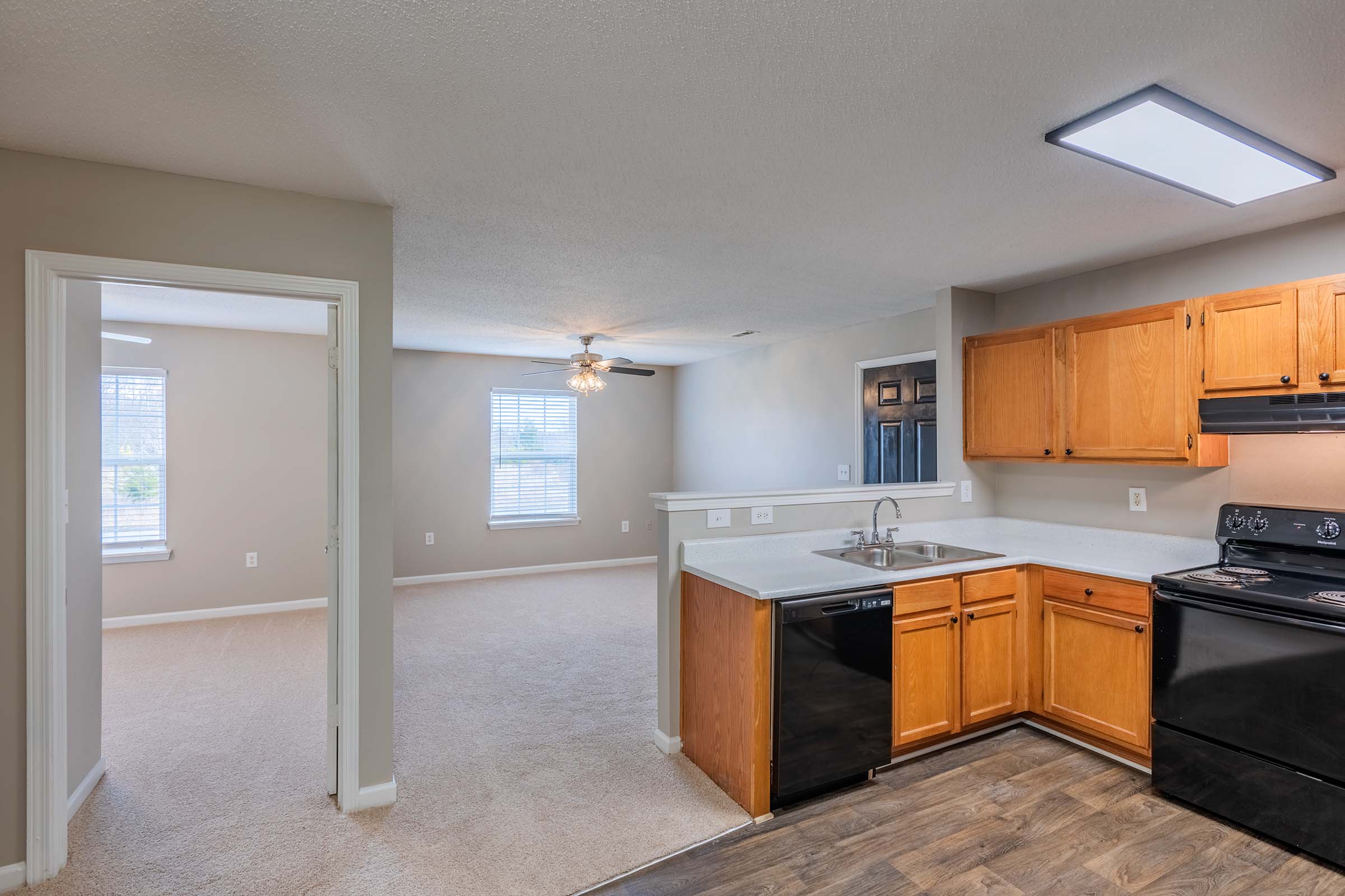 View of a modern kitchen and living area. The kitchen features wooden cabinets, a black dishwasher, and stove. Adjacent to the kitchen is a spacious living room with light-colored carpeting, a ceiling fan, and large windows, providing natural light. The walls are painted in a neutral tone.