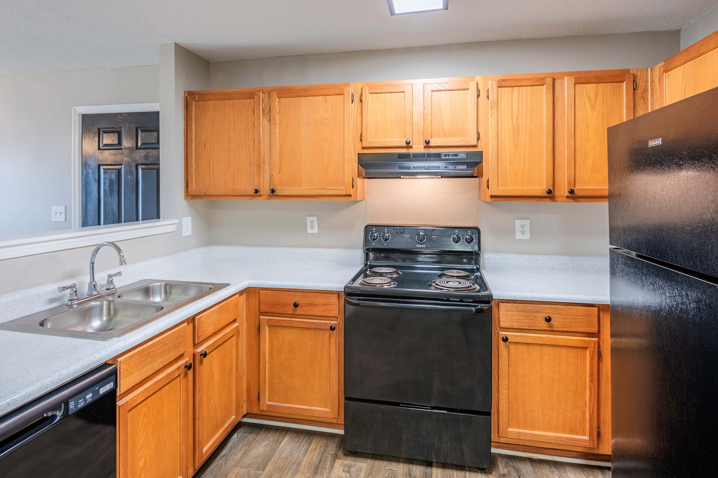 Modern kitchen featuring wooden cabinets, a black stove and refrigerator, stainless steel sink, and a countertop with a light-colored surface. The space is well-lit with a ceiling light, and there are dark doors visible in the background, adding to the contemporary aesthetic.