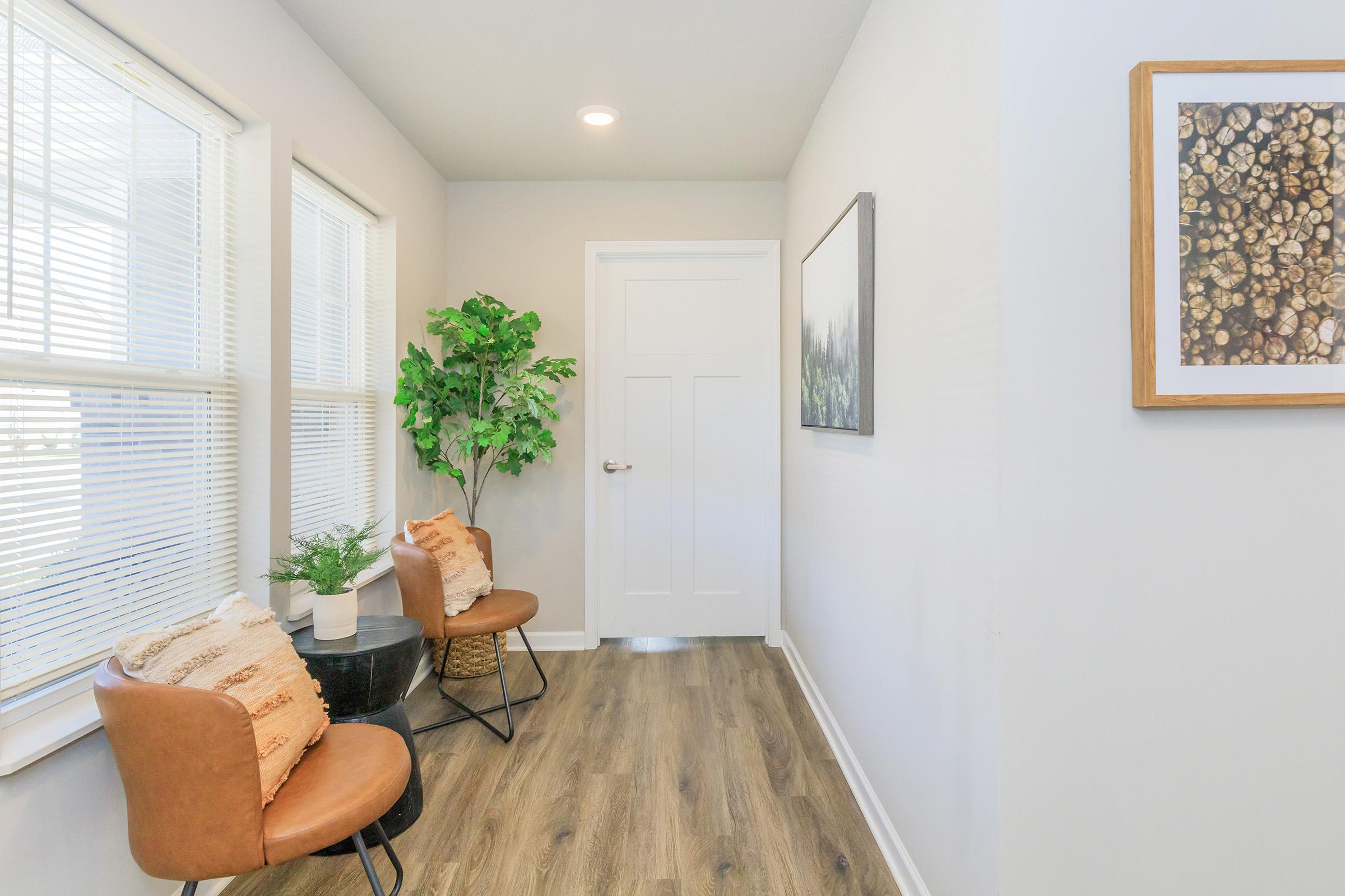 A well-lit hallway featuring two brown chairs and a round side table. A potted plant adds a touch of greenery, while framed artwork adorns the wall. The floor is made of wooden planks, and there's a white door at the end of the hallway. Natural light streams in through the windows, enhancing the space's inviting atmosphere.