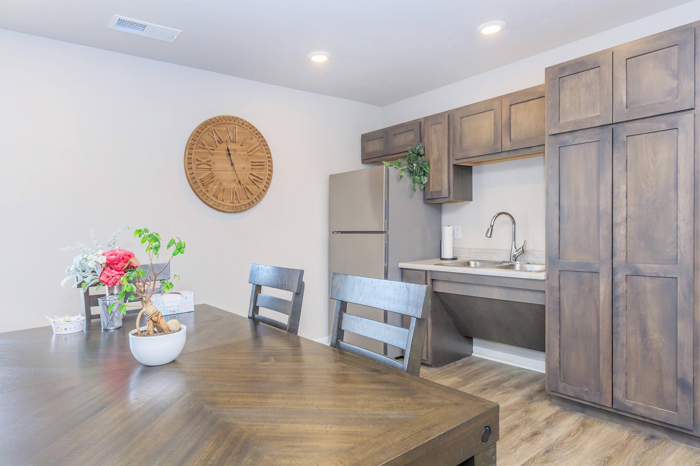 A modern kitchen area featuring a wooden dining table with chairs, a large clock on the wall, a stainless steel refrigerator, and a sink. The kitchen cabinetry is dark wood, and there are decorative plants and a flower arrangement on the table, creating a warm, inviting atmosphere.