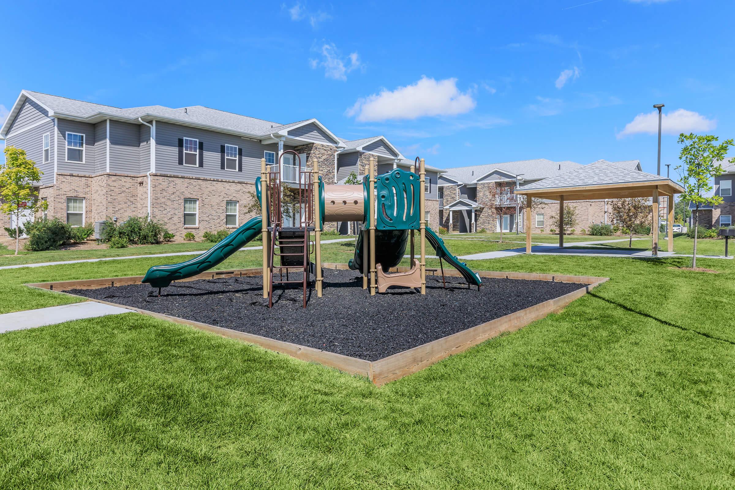 A playground featuring a slide and climbing structure, surrounded by green grass and black rubber mulch, with nearby residential buildings and a shaded seating area. The sky is clear with a few clouds, creating a bright and inviting atmosphere.