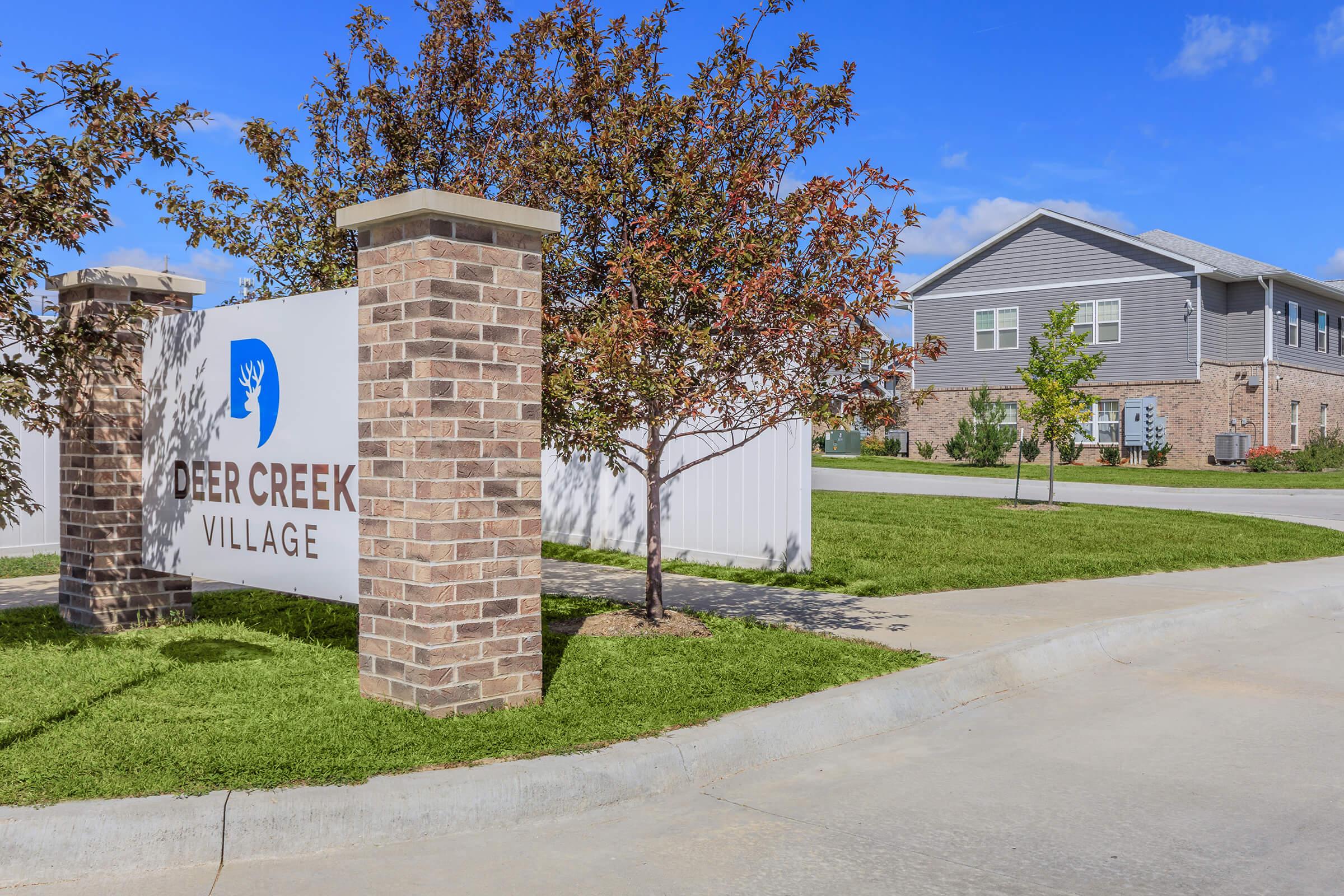 Sign for Deer Creek Village with brick pillars, surrounded by green grass and trees. In the background, there is a building with a gray exterior and a blue sky with clouds. The entrance is well-maintained, suggesting a welcoming residential community.