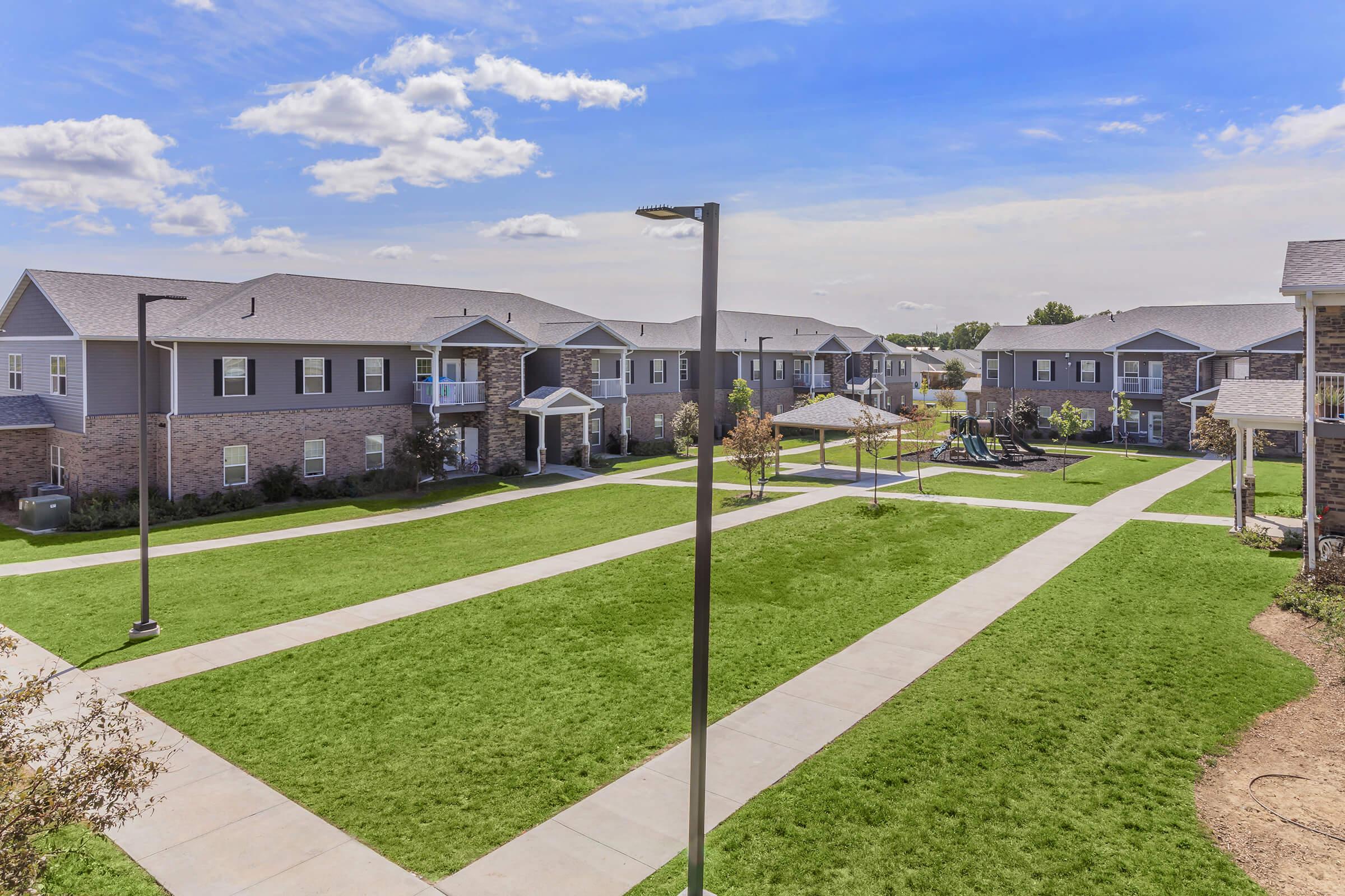 A spacious residential community with well-maintained grass lawns and sidewalks. The area features apartment buildings, trees, and a playground in the background under a clear blue sky. Several lampposts line the paths, creating an inviting atmosphere for residents and visitors.
