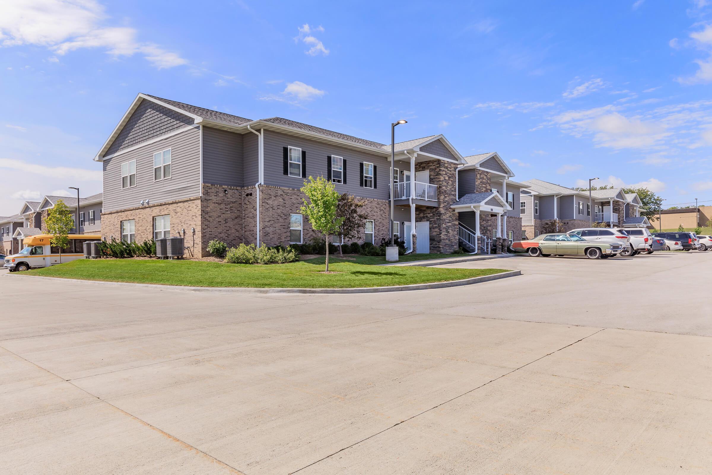 A modern apartment complex featuring two-story buildings with a mix of brick and siding exteriors. The well-kept lawn is surrounded by parking spaces, with various vehicles parked nearby. The sky is clear with a few clouds, contributing to a bright and welcoming atmosphere.