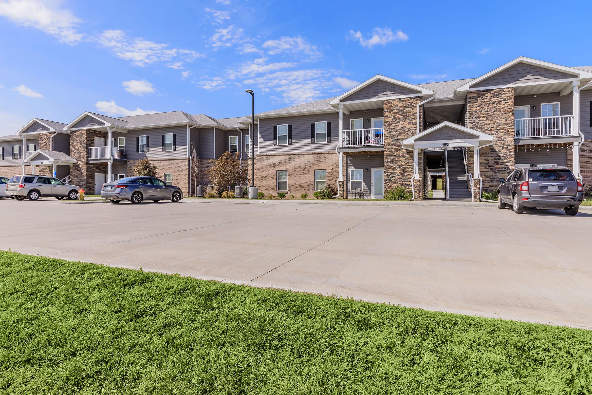 Two-story apartment building with stone accents and a row of parked cars in front. The entrance features balconies and a landscaped lawn. The sky is clear with a few clouds, giving a bright and welcoming atmosphere.