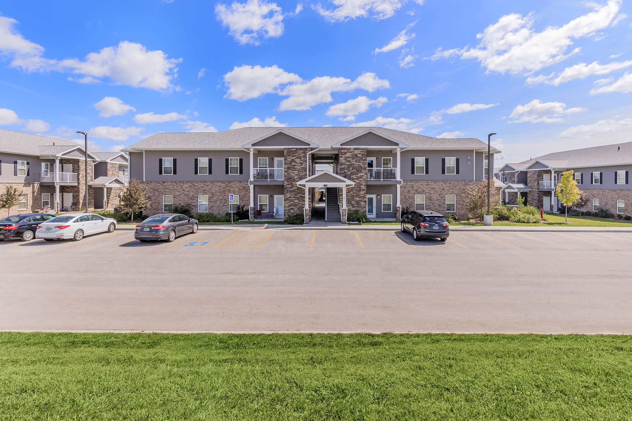 A modern apartment complex with multiple units, featuring a combination of stone and siding exteriors. The scene includes a landscaped area with grass in front, several parked cars along a paved parking lot, and a clear blue sky with fluffy white clouds above.