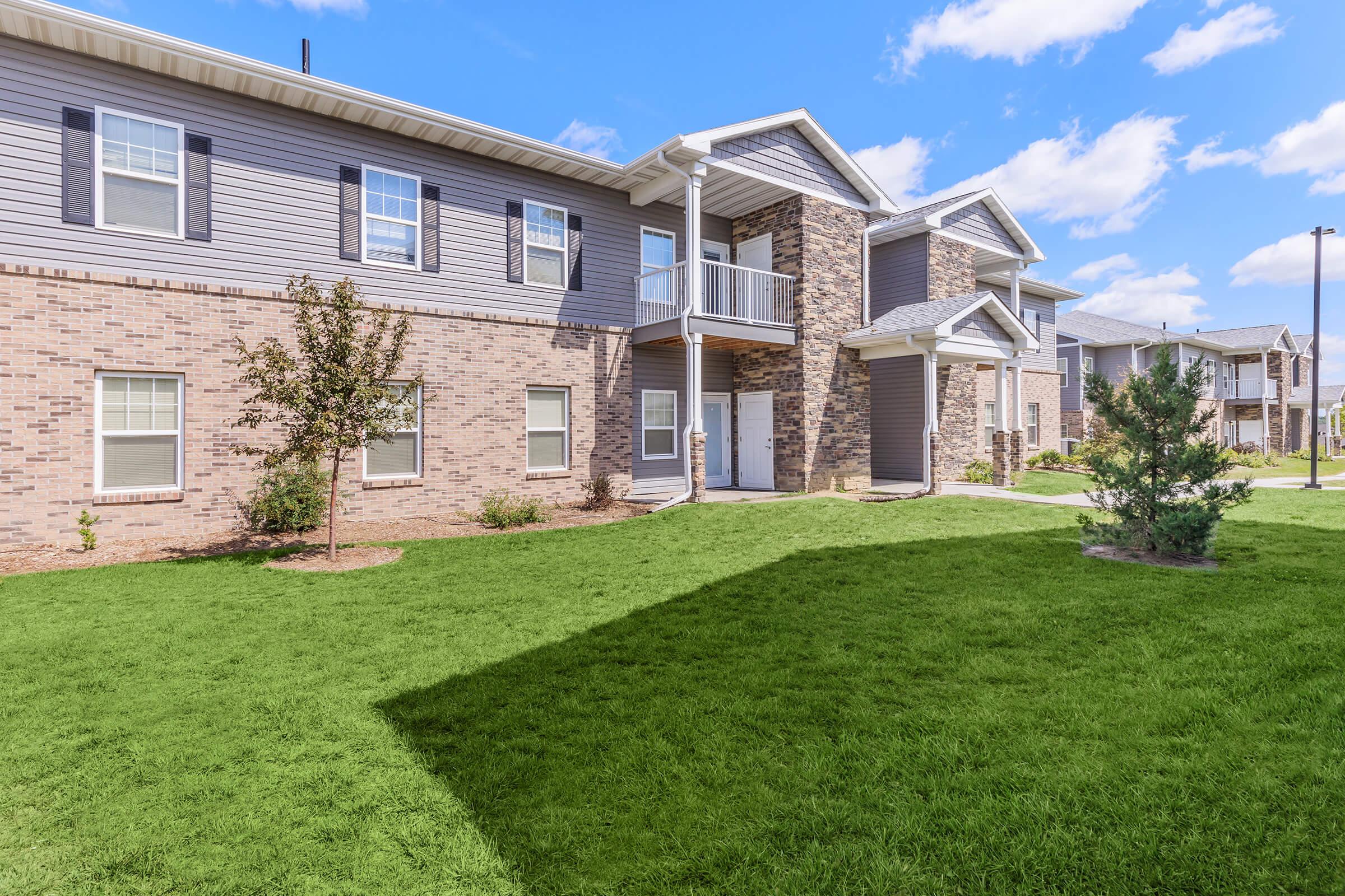 Two-story apartment building with a mix of brick and siding exterior. The building features several windows and a balcony on the second floor. Lush green lawn surrounds the building, with small trees and landscaping. The sky is partly cloudy with blue hues.