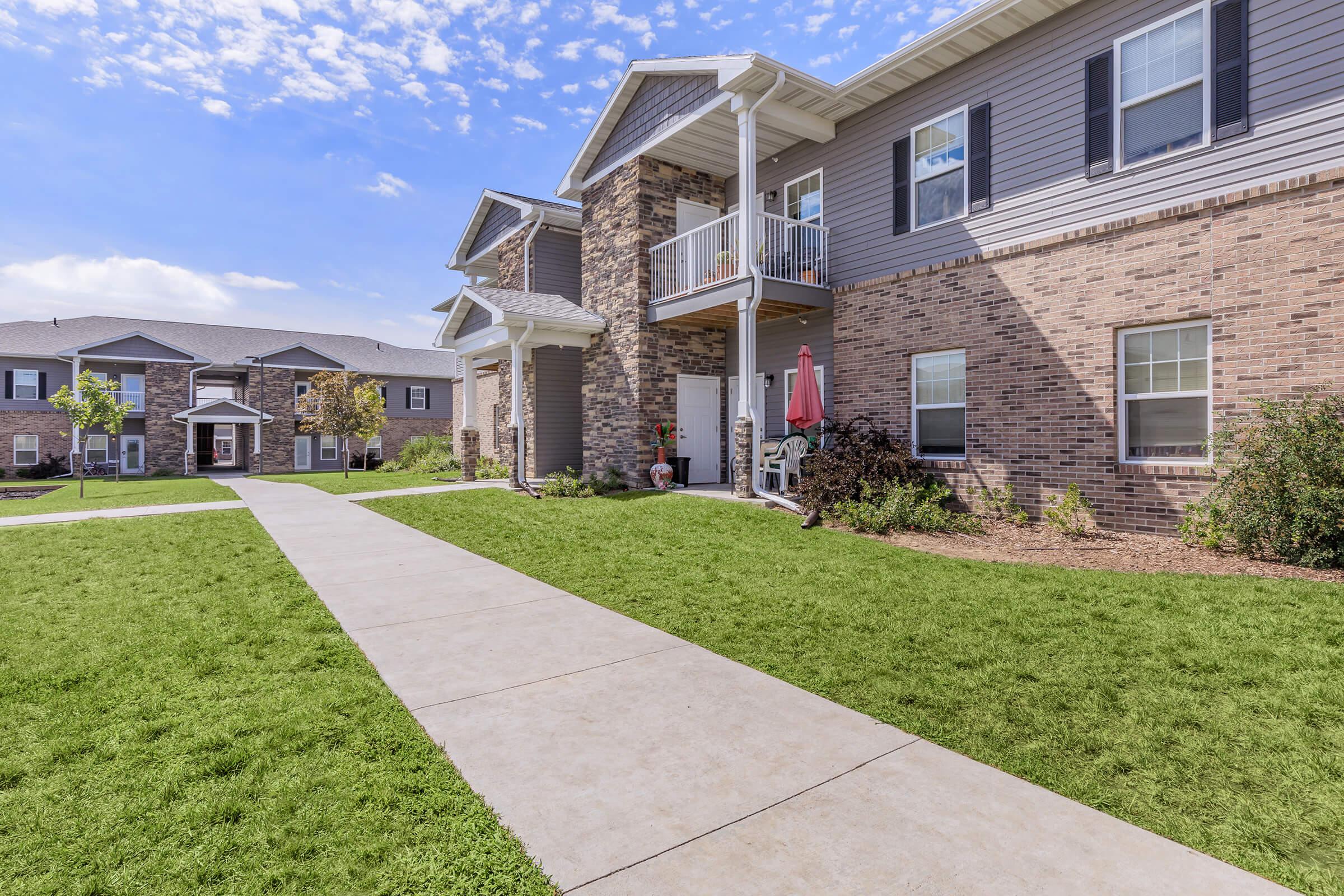 A view of a residential building with multiple units, featuring a well-maintained lawn and pathway. The architecture includes brick and siding, with balconies and outdoor seating. Clear blue sky and greenery enhance the inviting atmosphere of the community.