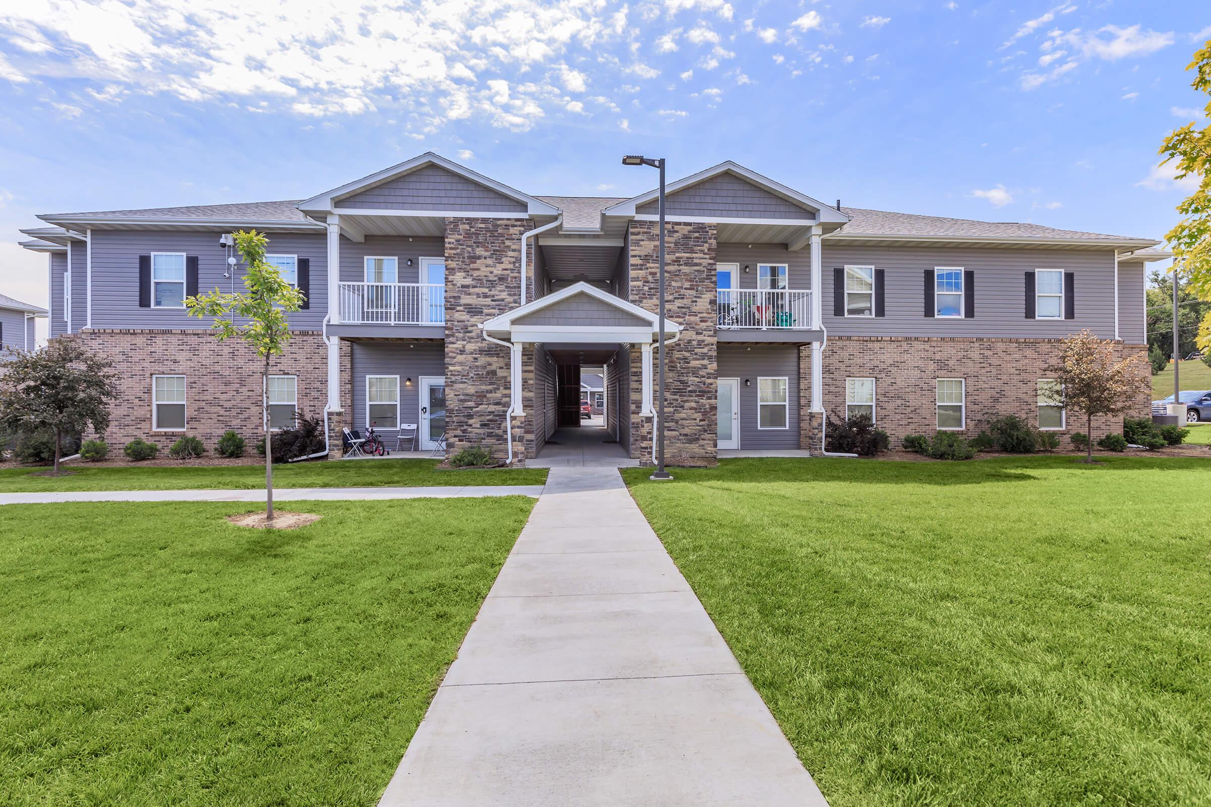 A modern two-story apartment building with a combination of stone and siding exteriors, featuring a central entrance, balconies, and well-maintained landscaping. A paved walkway leads to the entrance, surrounded by green grass and small trees. Bright sky with scattered clouds in the background.