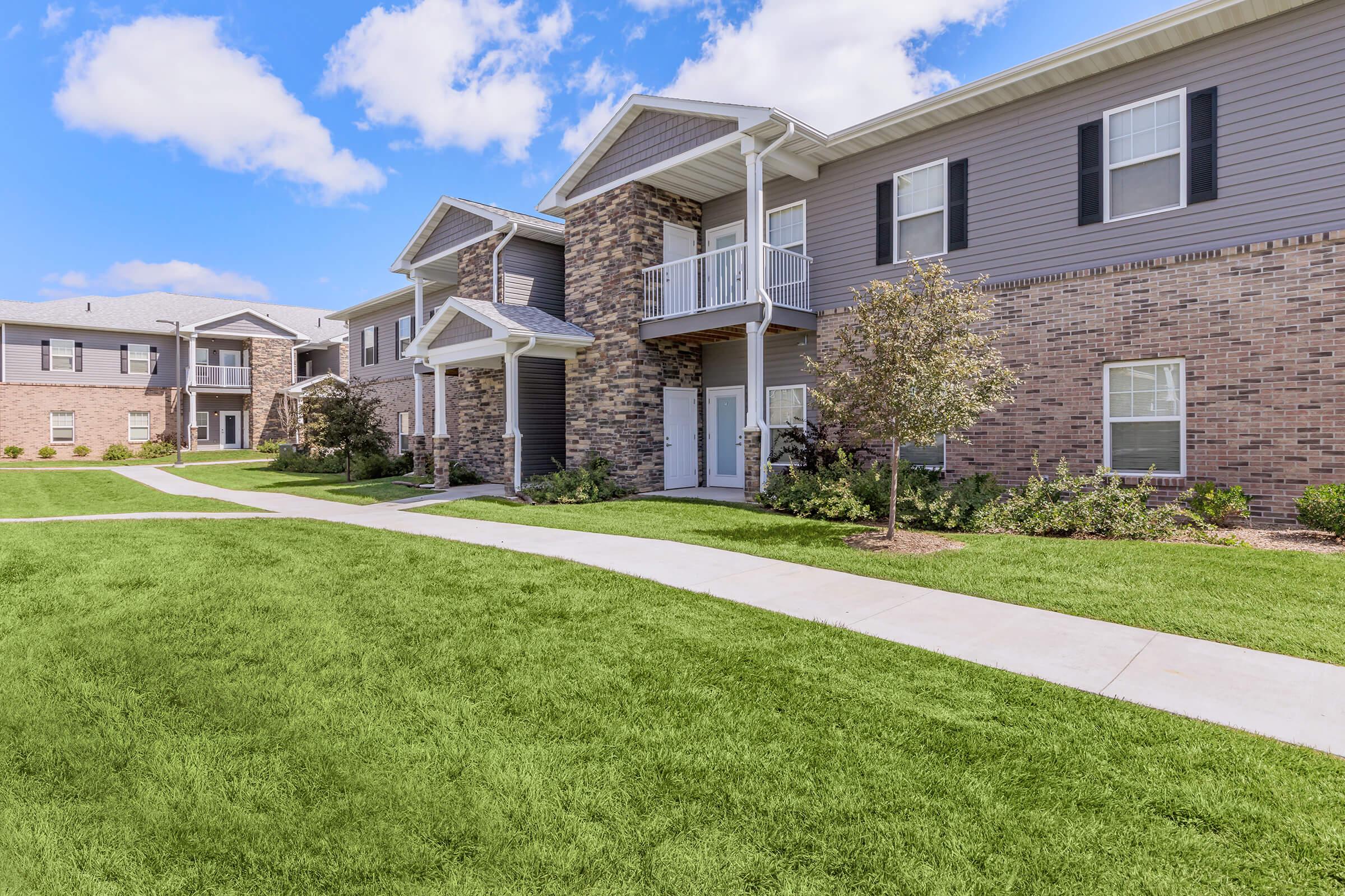 Two-story apartment buildings with a mix of brick and siding exteriors, featuring balconies and well-maintained landscaping. A wide, paved walkway meanders through green grass, connecting the units. The sky is bright and partly cloudy, enhancing the serene atmosphere of the residential area.