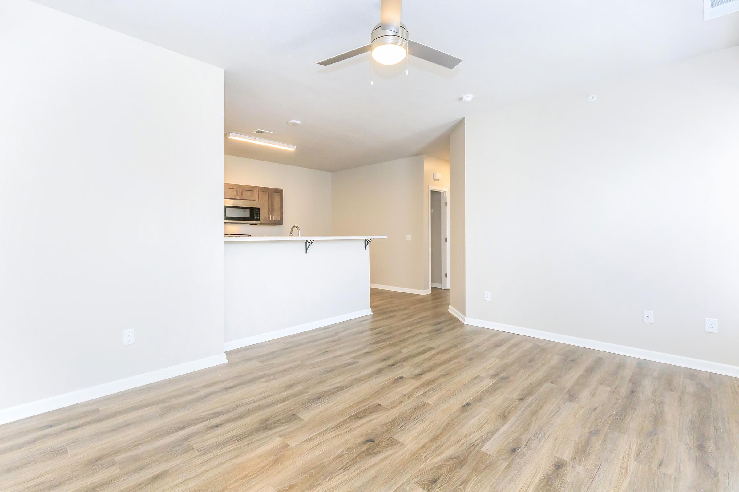 A spacious, modern living area with light-colored walls and wooden flooring. The room features a ceiling fan and an open layout leading to a kitchen with stainless steel appliances. An adjacent doorway leads to another room. Natural light brightens the space, creating a welcoming atmosphere.