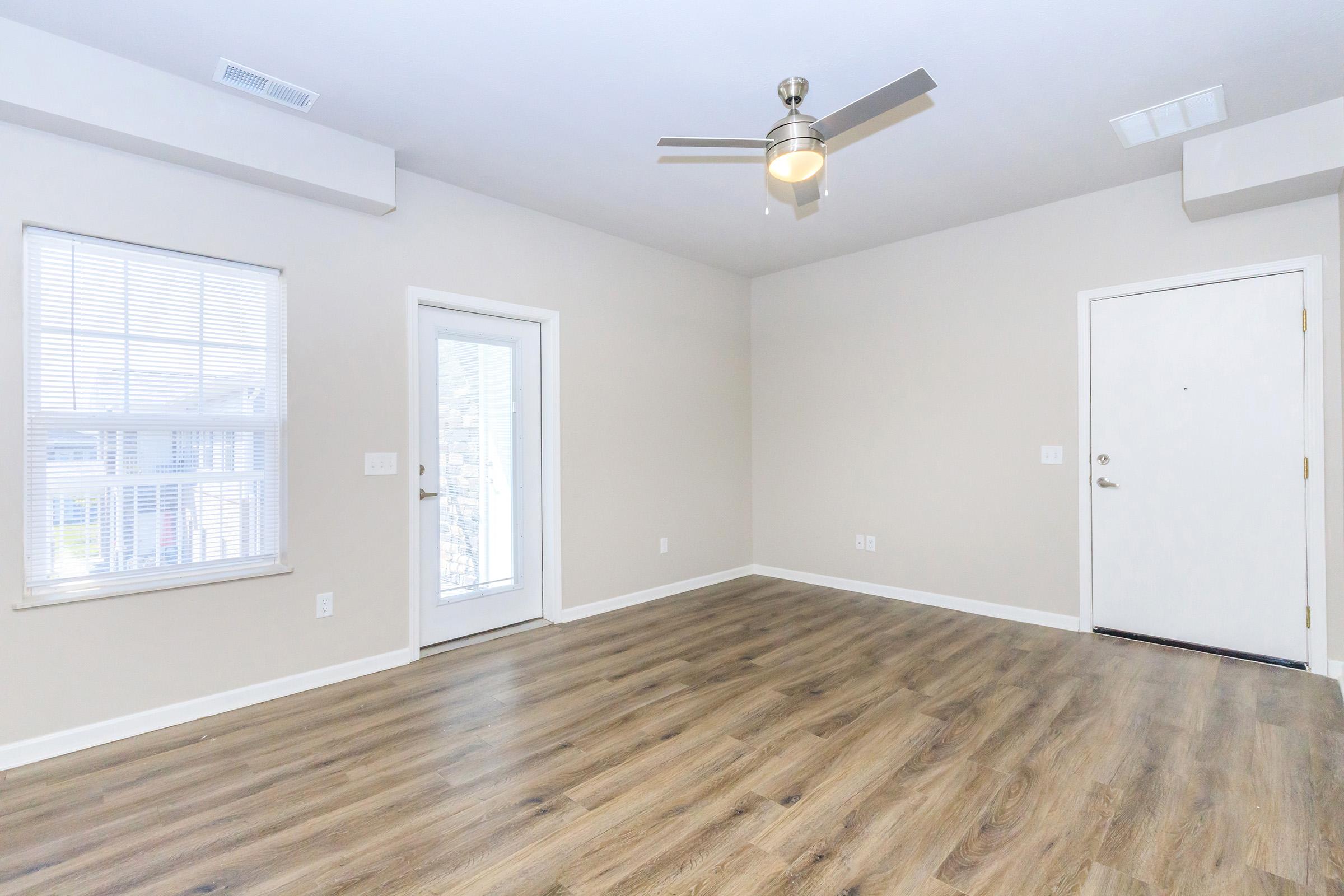 A well-lit, empty room with light beige walls and hardwood flooring. It features a ceiling fan, a door to the outside, and a white-paneled door leading to another room. Large windows allow natural light to fill the space, creating an airy and open atmosphere.
